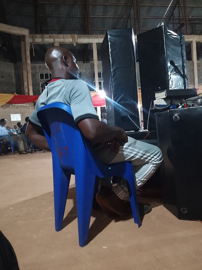 A bald man sits on a blue plastic chair, wearing a grey shirt and striped pants. He is positioned sideways, looking towards two tall black speakers. In the background, several people are seated in chairs and there's a glimpse of red and yellow fabric. The setting appears to be an indoor space, possibly a warehouse or large hall with an exposed ceiling structure. A sound system is set up near the speakers.