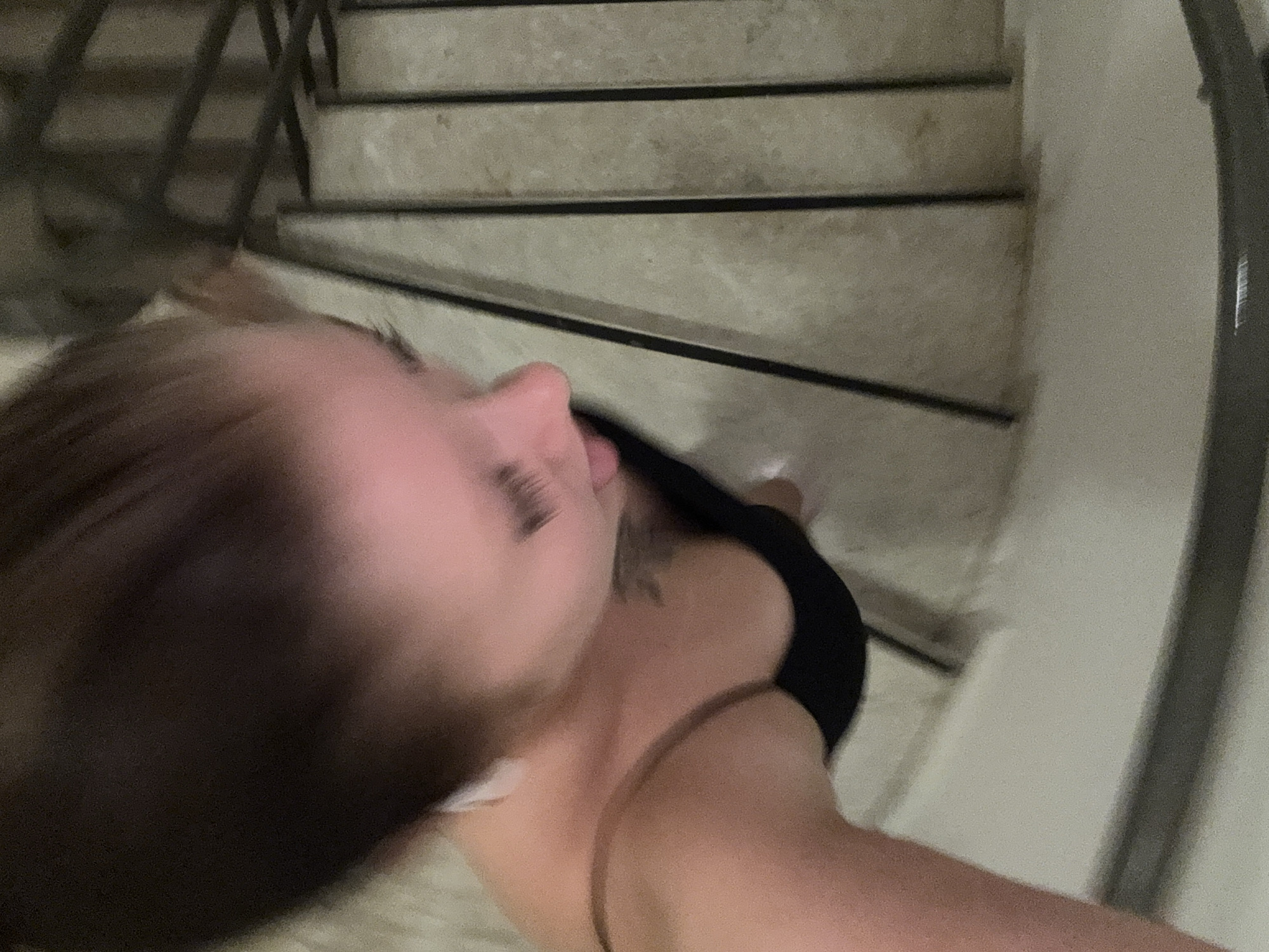 The image shows a close-up, blurry shot of a person's face and upper body. The person has dark hair pulled back and is wearing a black top. They appear to be indoors, possibly on a staircase, with a textured floor and metal railings visible. The image is overexposed in some areas, creating a pinkish hue on the skin. There is a tattoo visible on the person's chest. The location is identified as Aksu, Turkey.