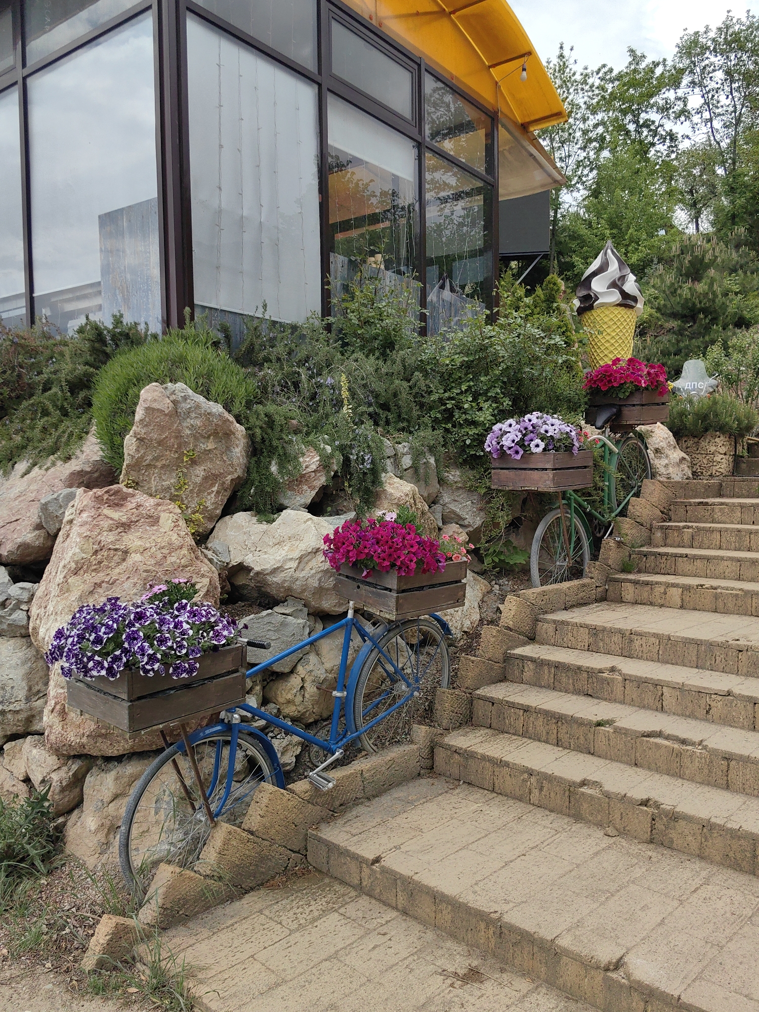 The scene depicts an outdoor display in Luchistoe, Ukraine. Two bicycles, one blue and one green, are positioned beside a set of stone stairs. Each bicycle has a wooden planter box attached, filled with colorful flowers. A large, simulated ice cream cone stands behind the green bicycle. The bicycles and stairs are surrounded by rocks, greenery, and a building with large windows and a yellow awning. The sky appears overcast.