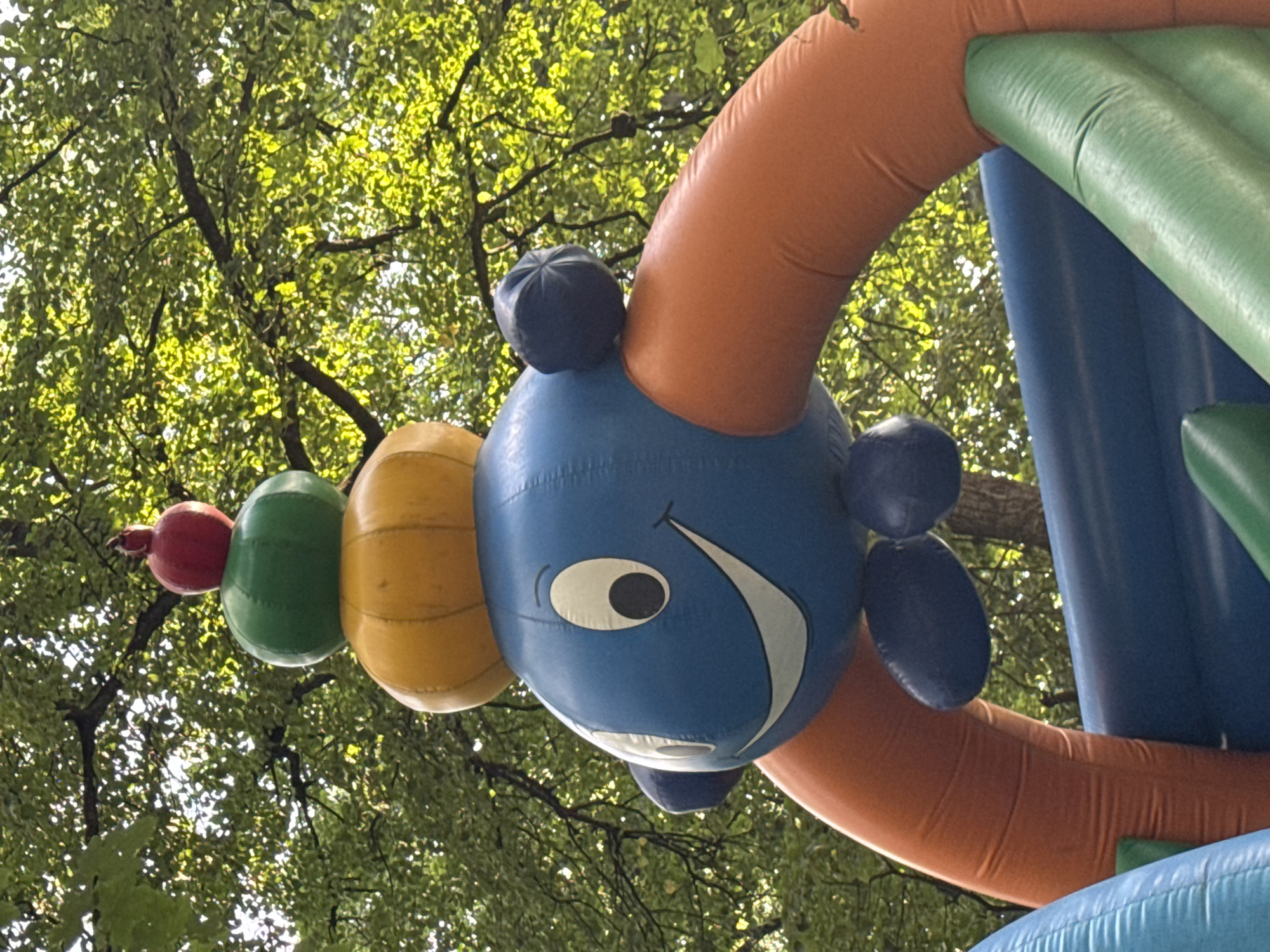 This is a close-up, eye-level, outdoor, medium shot of a portion of a blue inflatable bounce house. The bounce house is decorated with an arrangement of stacked, balloon-like shapes on top, resembling fruit. The topmost element is a small, red, pomegranate-shaped balloon. Below it is a green, round balloon, and beneath that is a larger, yellow, pumpkin-shaped balloon. The main body of the bounce house is blue and features a cartoonish face with large oval white eyes, black pupils, and a wide white smiling mouth. Two small, dark blue, puffed-out shapes, resembling ears or cheeks, are attached to the sides of the blue head. Orange, inflatable arched structures form part of the bounce house's frame, curving around the blue head. The background consists of lush green tree foliage, suggesting the bounce house is located outdoors, possibly in a park or backyard in Berezhany, Ukraine. The lighting indicates it is daytime, with sunlight filtering through the leaves. No people are visible in the image.