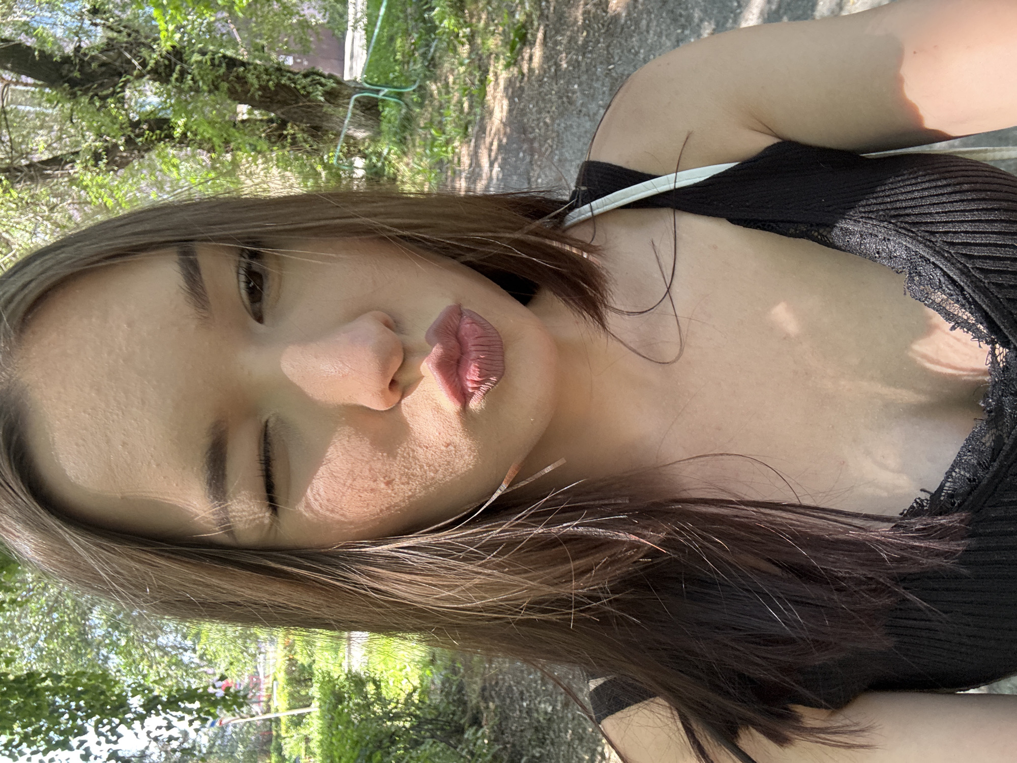 A young woman with long, dark hair and fair skin winks at the camera, pursing her lips. She is outdoors, possibly in a park, with lush green trees and foliage behind her. The sunlight casts dappled shadows on her face and shoulders. She is wearing a black tank top with lace trim. The background suggests a sunny day in a relatively unpopulated area. The image is a close-up, focusing on the woman's face and upper body. No specific landmarks in Belovo, Russia, are visible.