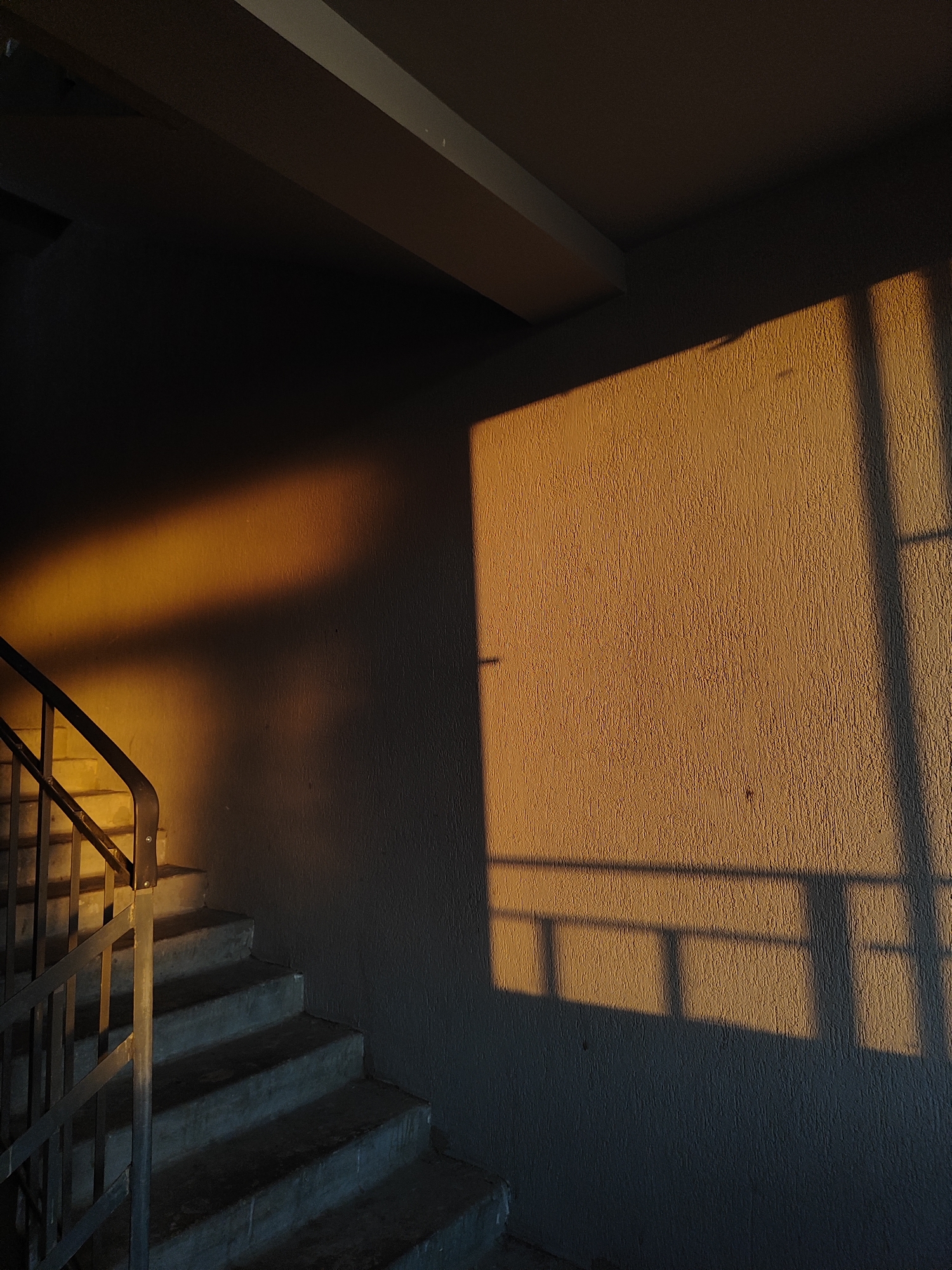 The image captures an indoor stairwell bathed in warm, golden light, suggesting either early morning or late afternoon (golden hour). On the left, a concrete staircase with a dark metal railing ascends from the bottom left corner. Parts of the stairs and the wall directly adjacent to them are illuminated by a soft, diffused golden light.
The majority of the right side of the frame is dominated by a textured, light brown or beige wall. A large, rectangular patch of intense golden sunlight illuminates a significant portion of this wall. Within this illuminated area, distinct dark shadows are cast, resembling the grid or frame of a window. Another diagonal strip of golden light extends across the upper left wall, creating a stark contrast with the unlit, darker sections.
Overhead, parts of a ceiling and structural beams are visible, mostly in shadow, with a thin line of light highlighting one edge of a beam at the very top. The shadows throughout the scene are sharp and well-defined. The overall setting is a functional interior space, possibly a common area in a building like an apartment complex or industrial facility. There are no people present, no activity is taking place, and no visible text.