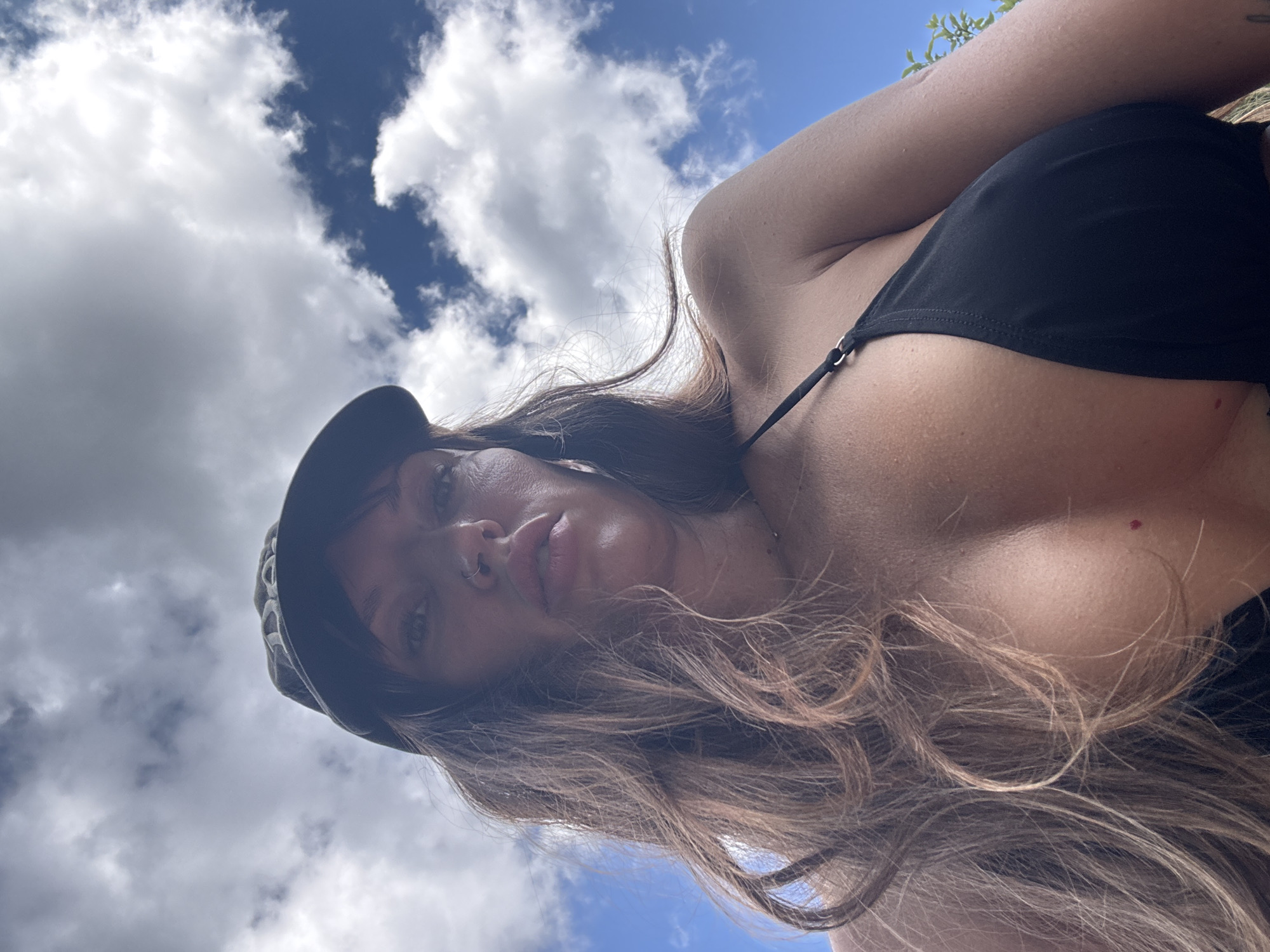 The original image, and its crops, depict a low-angle, close-up shot of a woman from the shoulders up, against a backdrop of a blue sky with white clouds. She is wearing a black bikini top and a dark baseball cap. Her long, brown hair is flowing. Her face is angled downwards, looking towards the camera. She has a nose ring and appears to be wearing light makeup. The setting is outdoors, under a bright sky. There is no indication or visual information within the image that would allow for the identification of the specific location as Красная Пресня Снт, Russia.