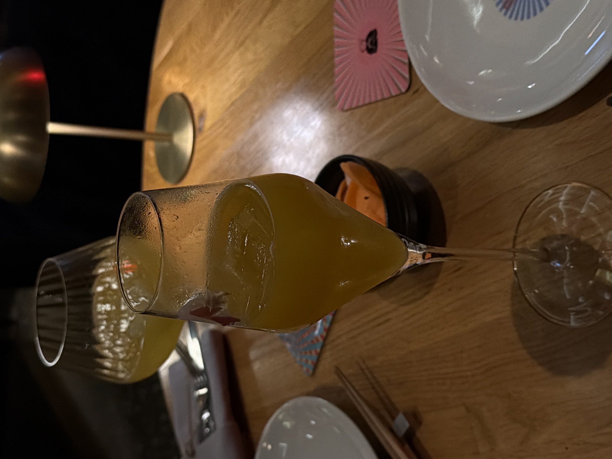 A high-angle shot captures a dimly lit dining table scene, potentially in Belgrade, Serbia. Two champagne glasses are present, one filled with an orange beverage and ice cubes, lying horizontally, and the other glass is empty. Surrounding these are a small black bowl containing orange slices, a white plate with chopsticks, a pink and white coaster, and a lamp with a reflective shade. The environment suggests a restaurant or bar setting.