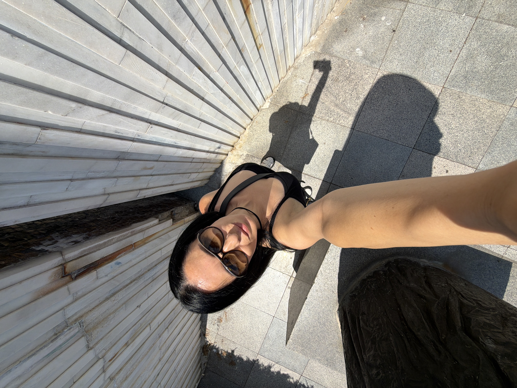 A woman with black hair and sunglasses is taking a selfie in a sunny outdoor area. She is wearing a black tank top. The background consists of a textured wall with horizontal panels and a tiled ground. A shadow of the woman is cast on the ground. There is a dark, textured object in the lower left of the frame. The scene appears to be in the city of Pleven, Bulgaria.