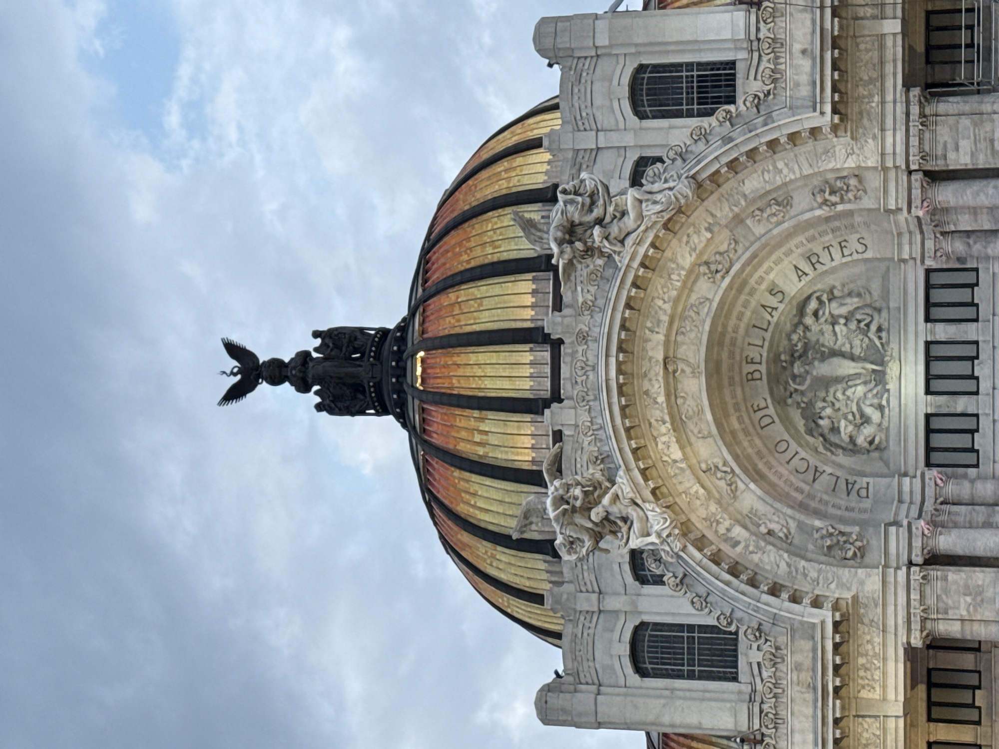 This image shows the Palacio de Bellas Artes in Mexico City, Mexico. The shot is taken from a low angle, capturing the dome of the palace and a portion of the facade. The dome is striped with alternating bands of gold and dark metal. Atop the dome sits a dark sculpture. The facade is made of gray stone and adorned with sculptures. The phrase "Palacio de Bellas Artes" is visible, carved into the stone. The sky behind the palace is cloudy.