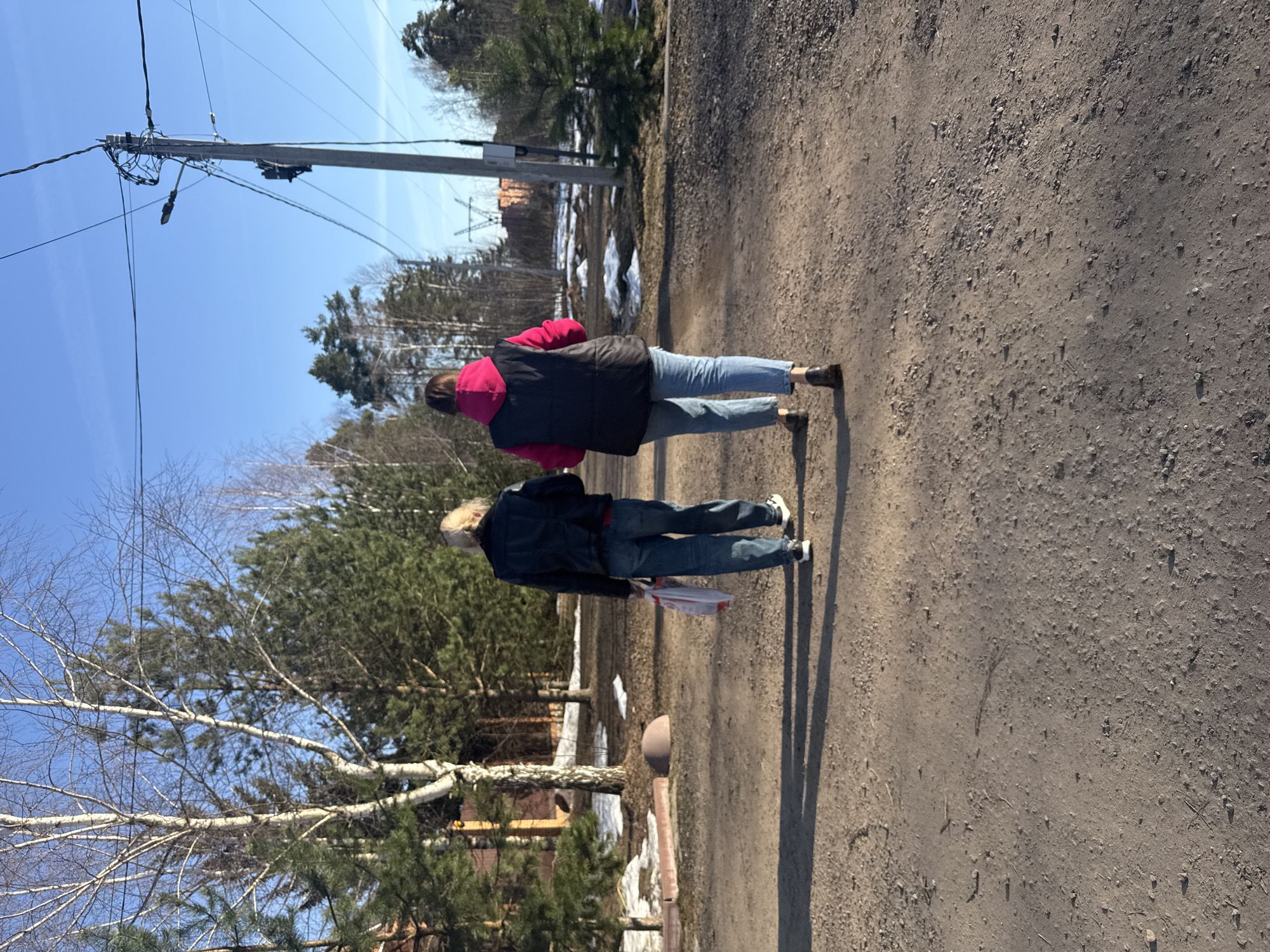 The image shows two people walking on a dirt road in a rural area near Dmitrov, Russia. The woman in front is wearing a pink jacket with a black vest and jeans, while the person behind her is wearing a dark jacket and jeans, and appears to be holding a white bag. The road is unpaved and dusty. On either side of the road are trees and patches of melting snow, suggesting it is early spring. A power pole is visible in the background against a clear blue sky.
