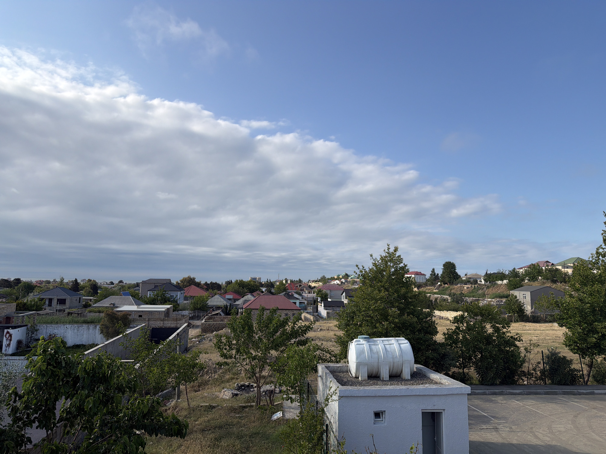 The image depicts a residential scene in Novxanı, Azerbaijan, under a sky partially covered with clouds. A small white building with a water tank on its roof dominates the foreground. Beyond the building, there are trees and other vegetation in a yard. Houses of varying designs, many with red roofs, are visible in the middle ground. In the background, there is a suggestion of more buildings stretching toward the horizon. The atmosphere appears calm and peaceful.