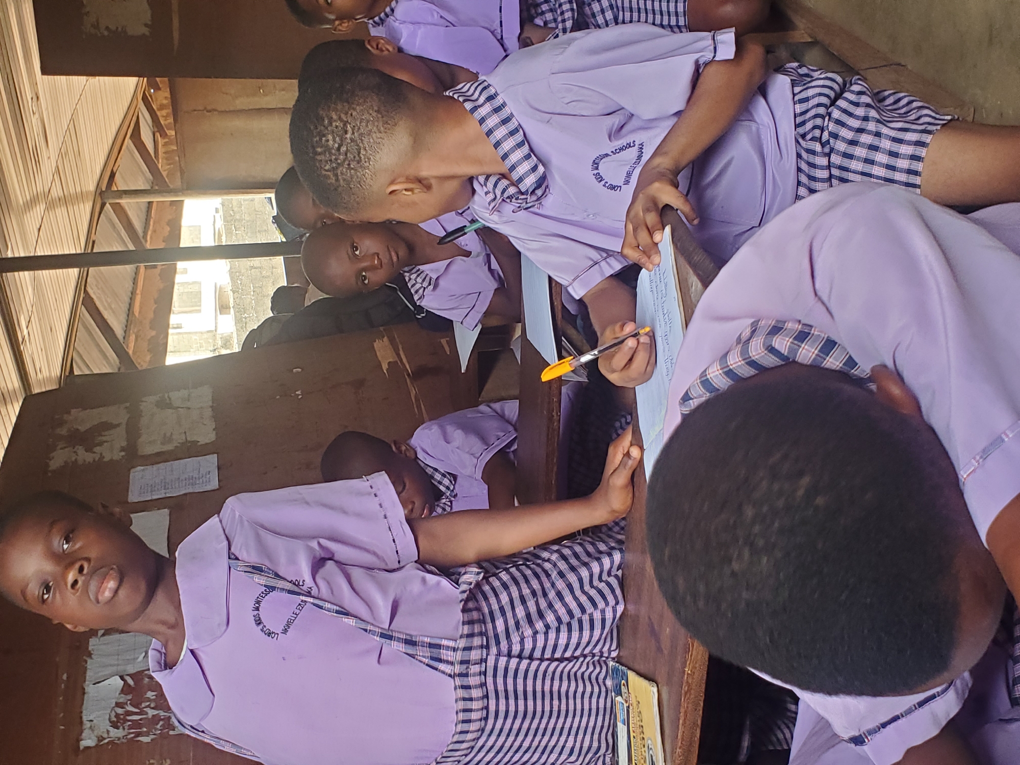 In a brightly lit room in Ukwu-Abwa, Nigeria, several children are seated at wooden desks, seemingly engaged in a learning activity. They are dressed in lavender shirts with plaid collars and plaid shorts/skirts. One child is writing on a piece of paper with an orange pen, while another is looking towards him. There are other children in the background, focused on their own tasks. The room has a partially visible corrugated metal ceiling. The walls are bare except for some papers affixed to them.