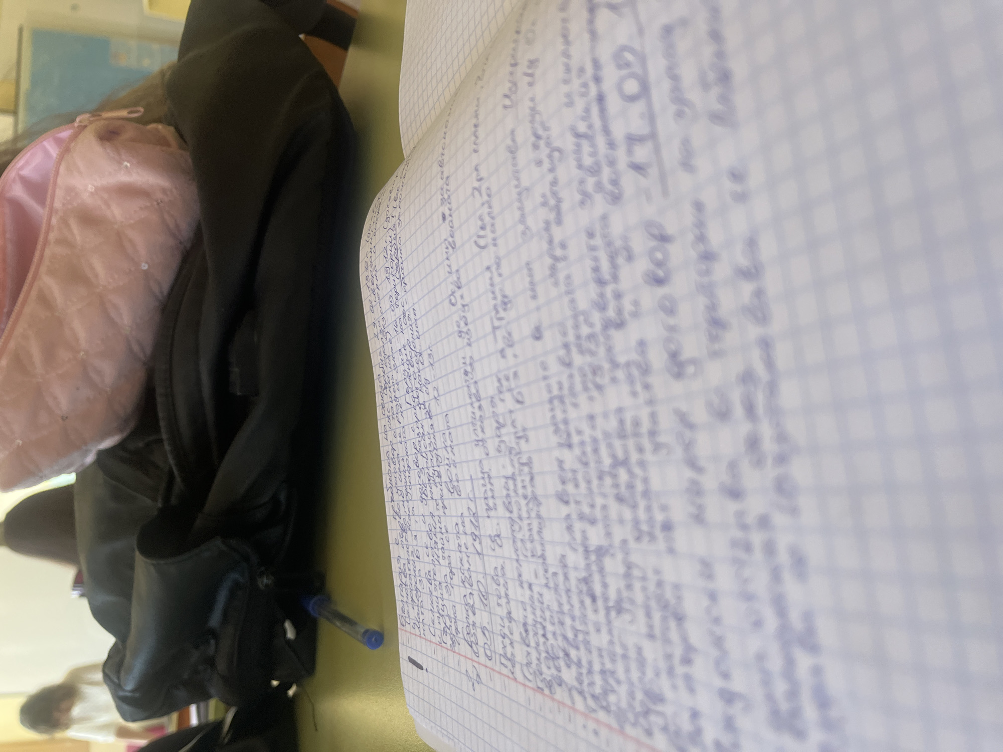The image shows a notebook with handwritten notes in a language likely Bulgarian. The notebook is resting on a desk or table, and nearby are items including a pink bag, a black jacket, and a pen. A person is visible out of focus in the background. The date "17.09" is written on the page. The location cannot be determined from the image.