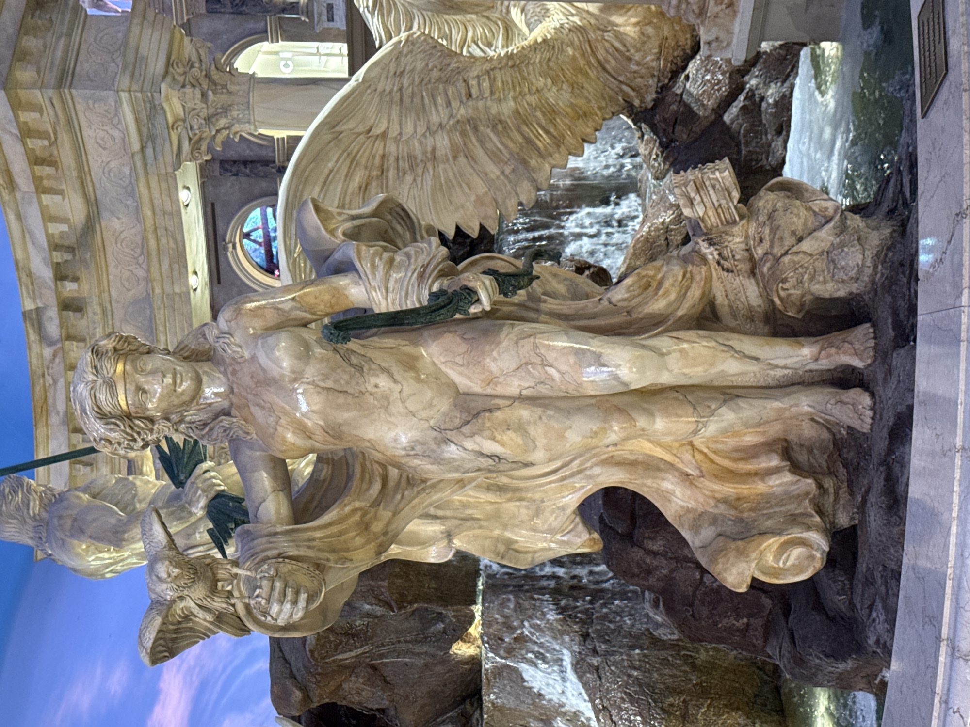The image captures a detailed view of a large, ornate fountain, likely a centerpiece in a grand interior setting. The fountain features multiple sculpted figures, prominently a central female statue made of light-colored marble with visible veining. She is depicted in a dynamic pose, draped in flowing robes that reveal parts of her body. She holds a bird of prey, possibly an eagle, on her left hand and a green object, resembling a spear or scepter, in her right. Behind her, a larger, muscular male figure is partially visible, holding a similar green object.
The figures are integrated with a rocky landscape, from which water cascades down in several streams, creating a sense of movement and natural beauty. The water collects in a pool at the base of the fountain. Surrounding the fountain are architectural elements, including columns and arches, suggestive of classical or baroque design. The lighting in the scene is somewhat dim, with artificial lights illuminating the sculptures and creating highlights and shadows. The overall impression is one of opulence and artistic craftsmanship. The sky visible in the upper background appears to be a painted or simulated sky, suggesting an indoor environment designed to mimic an outdoor scene. A sign with the letters "CI" is visible in the background, potentially indicating a commercial establishment. The location is identified as Paradise, United States of America, which is likely a reference to an area within or near Las Vegas, Nevada, known for its extravagant resorts and casinos.