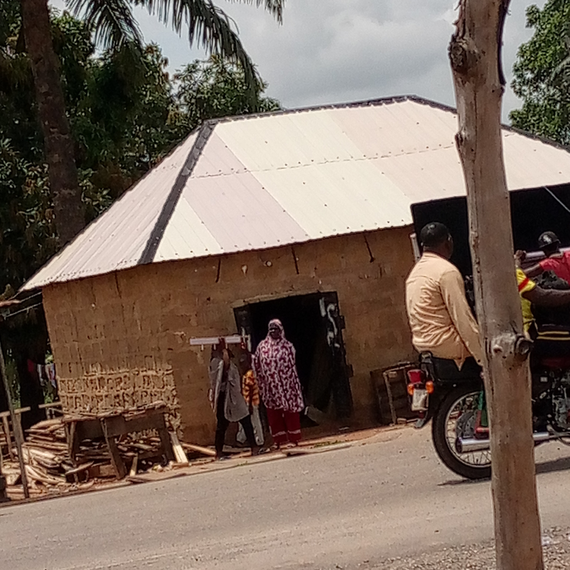 The image shows a street scene in Mararaba, Nigeria. A simple, square-shaped building with a white, corrugated metal roof stands to the left. The building appears to be constructed of mud bricks. In front of the building, there are three people and some lumber. A woman in a patterned, red and white robe stands in the doorway, while two other people stand to her left, one holding what looks like metal rods. To the right of the building, a man is seated on a motorcycle, partially obscured by a tree trunk in the foreground. The street is paved and there are trees in the background.