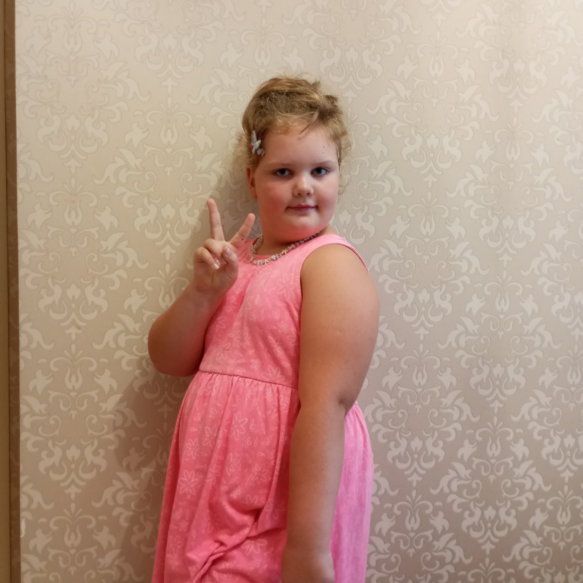 In Sochi, Russia, a young girl with blonde curly hair, adorned with a small silver clip, stands against a light-colored damask wallpaper. She is wearing a pink sleeveless dress with a floral pattern and a beaded necklace. The girl is looking towards the camera with a neutral expression and is making a peace sign with her right hand. Her left arm is bent at the elbow, resting by her side. The lighting appears to be artificial, creating soft shadows.