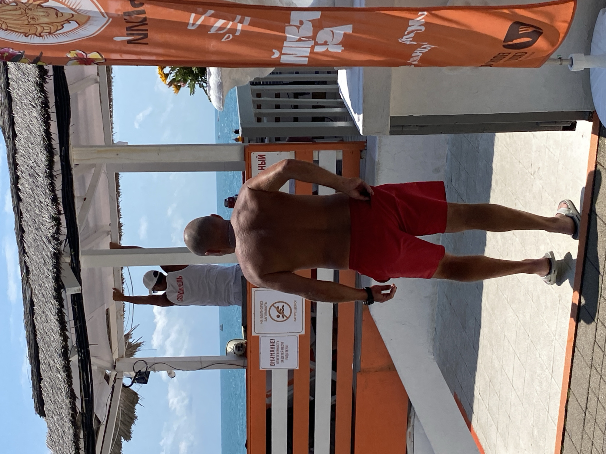The image shows two men near a beach in Sochi, Russia. One man, facing away from the camera, is wearing red shorts and is shirtless. He is standing on a tiled surface, with the sea and sky visible in the background. To his left, another man is wearing a white tank top with "СПАСАТЕЛЬ" (Rescuer) printed on it in red. He is wearing a white cap and is reaching up to a white structure.
Above the men, an orange banner with white lettering is partially visible. The banner features a stylized face and what appears to be text in a foreign language. There are also signs visible on an orange and white striped barrier behind the shirtless man. One sign has an icon of a person slipping and text that says "НА ВОЛНОРЕЗ ЗАПРЕЩЕНО" (Prohibited on the breakwater). Another sign, partially obscured, reads "ВНИМАНИЕ! ОТВЕТСТВЕННОСТЬ ЗА ДЕТЕЙ НЕСУТ РОДИТЕЛИ" (Attention! Parents are responsible for children).
The scene appears to be set during the daytime, with bright sunlight casting shadows. The weather seems to be pleasant, with a clear blue sky and a calm sea, suggesting a beach or resort environment. The overall atmosphere seems relaxed, typical of a sunny day at the coast.