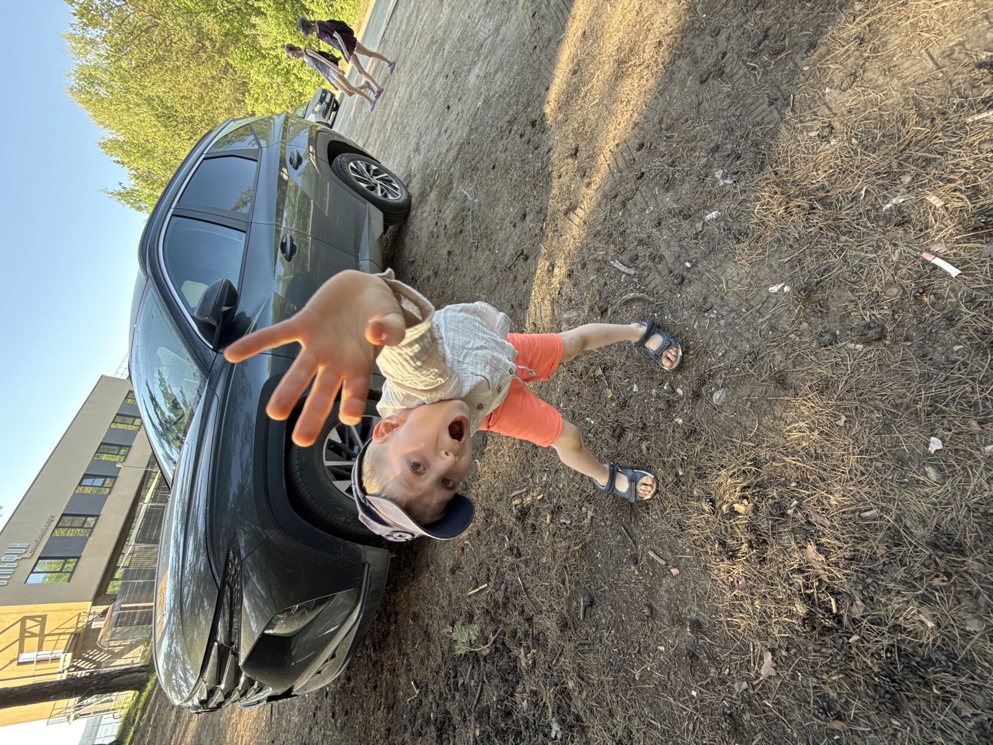 A young boy in orange shorts, a tan shirt, a baseball cap, and sandals stands on dirt and pine needles next to a gray Hyundai Tucson SUV. He is reaching out towards the camera. In the background, two figures are walking on a paved surface near green trees. A building is visible in the distance. The location is identifiable as the city of Chaikovsky, Russia.