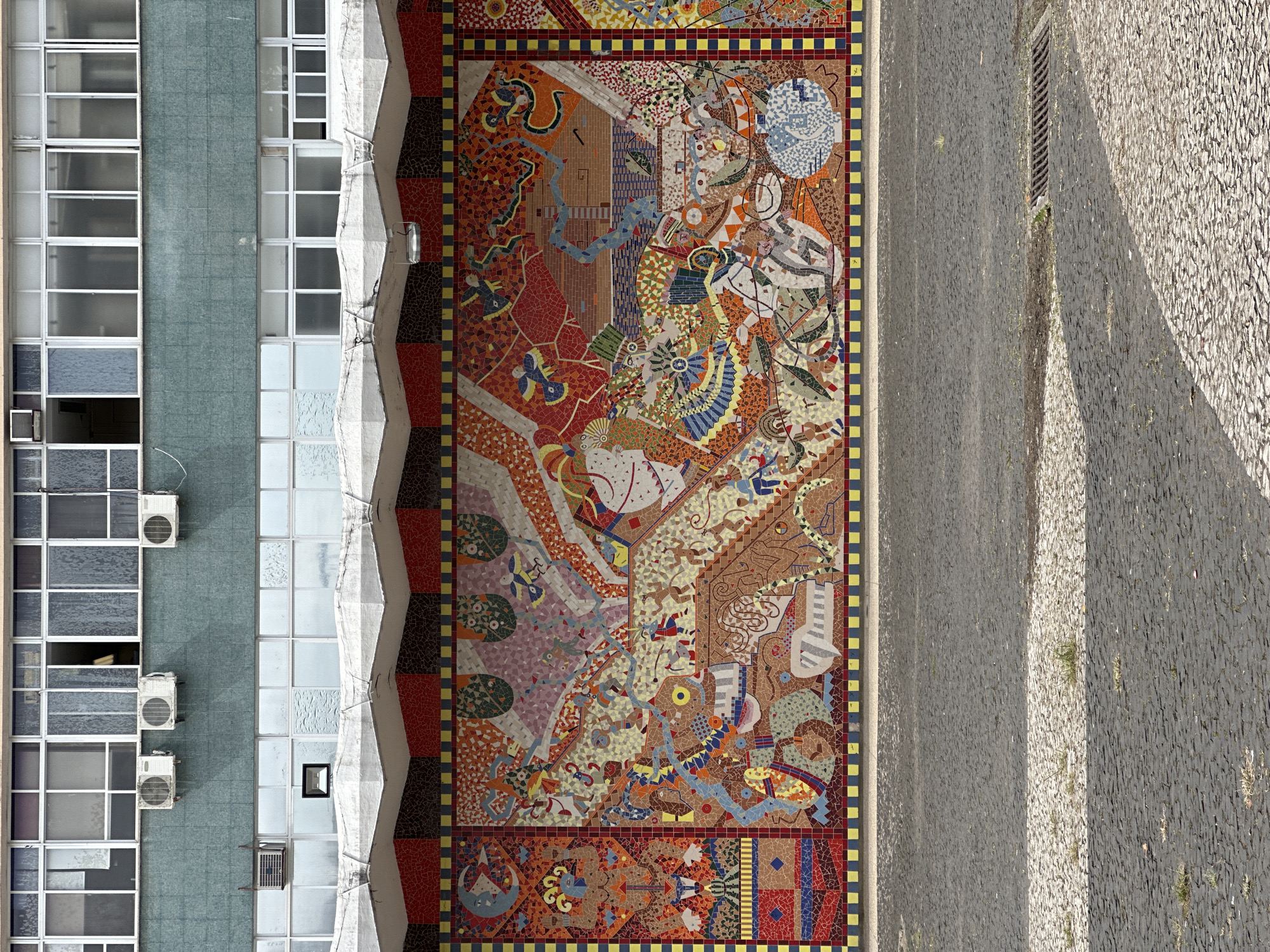 The image shows a large, colorful mosaic mural on the exterior wall of a building. The mural depicts various figures, symbols, and scenes, with a dominant palette of reds, oranges, yellows, blues, and greens. The mosaic is framed by a border of red and yellow squares.
Above the mural, the building features a series of windows and air conditioning units. The windows are divided into multiple panes and appear to be made of glass. The air conditioning units are white and mounted on the exterior wall. The building's facade is a mix of different materials and colors, including a teal-colored textured section between two bands of windows. The roofline of the building has a decorative, zigzagging white edge.
In the foreground, a cobblestone pavement stretches across the image. The pavement is made of dark gray stones with a lighter gray and white patterned strip running horizontally across it, possibly indicating a walkway or a drainage area. Sparse patches of grass and dirt are visible between the cobblestones. A storm drain grate is present in the pavement on the right side of the image.
The overall scene suggests an urban environment, likely a public space or the exterior of a public building. This mosaic is located in Florianópolis, Brazil, specifically on the walls of the Colégio Catarinense, a historic educational institution. The mural, created by artist Cláudio M. Linhares, is a significant piece of public art in the city.