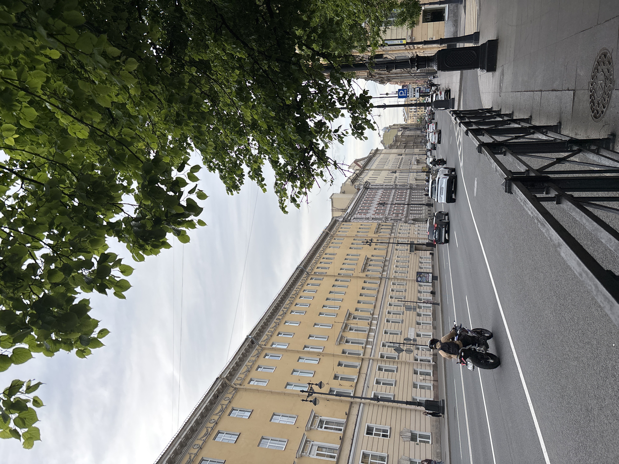 The image captures a rotated view of a wide city street in Saint Petersburg, Russia, under an overcast sky. The upper left portion of the frame is filled with a dense canopy of vibrant green leaves from a tree, suggesting a spring or summer setting.
The main subject is a multi-lane asphalt road, extending from the bottom to the top of the image (when viewed upright). White road markings delineate the lanes. A black metal fence with decorative elements runs along the right side of the road, separating it from the sidewalk. Traffic includes several cars, mostly light-colored, and a black motorcycle with two riders. The motorcycle, with its driver wearing a tan jacket and a helmet, is prominently featured in the mid-right.
To the right of the street, a long, multi-story building with a distinctive light yellow facade dominates the cityscape. It features numerous regularly spaced rectangular windows, decorative cornices, and architectural embellishments typical of historic European architecture. Another similarly styled building, possibly lighter in color, is visible next to it. Black lampposts line the street and sidewalk. The sidewalk itself is paved with light grey slabs, and a circular, patterned manhole cover is visible.
The sky appears overcast, indicating diffused lighting, and overhead power lines are visible against it. A blue "P" sign, likely indicating parking, can be seen further down the street. The scene depicts a typical urban environment with ongoing traffic and historical architecture.