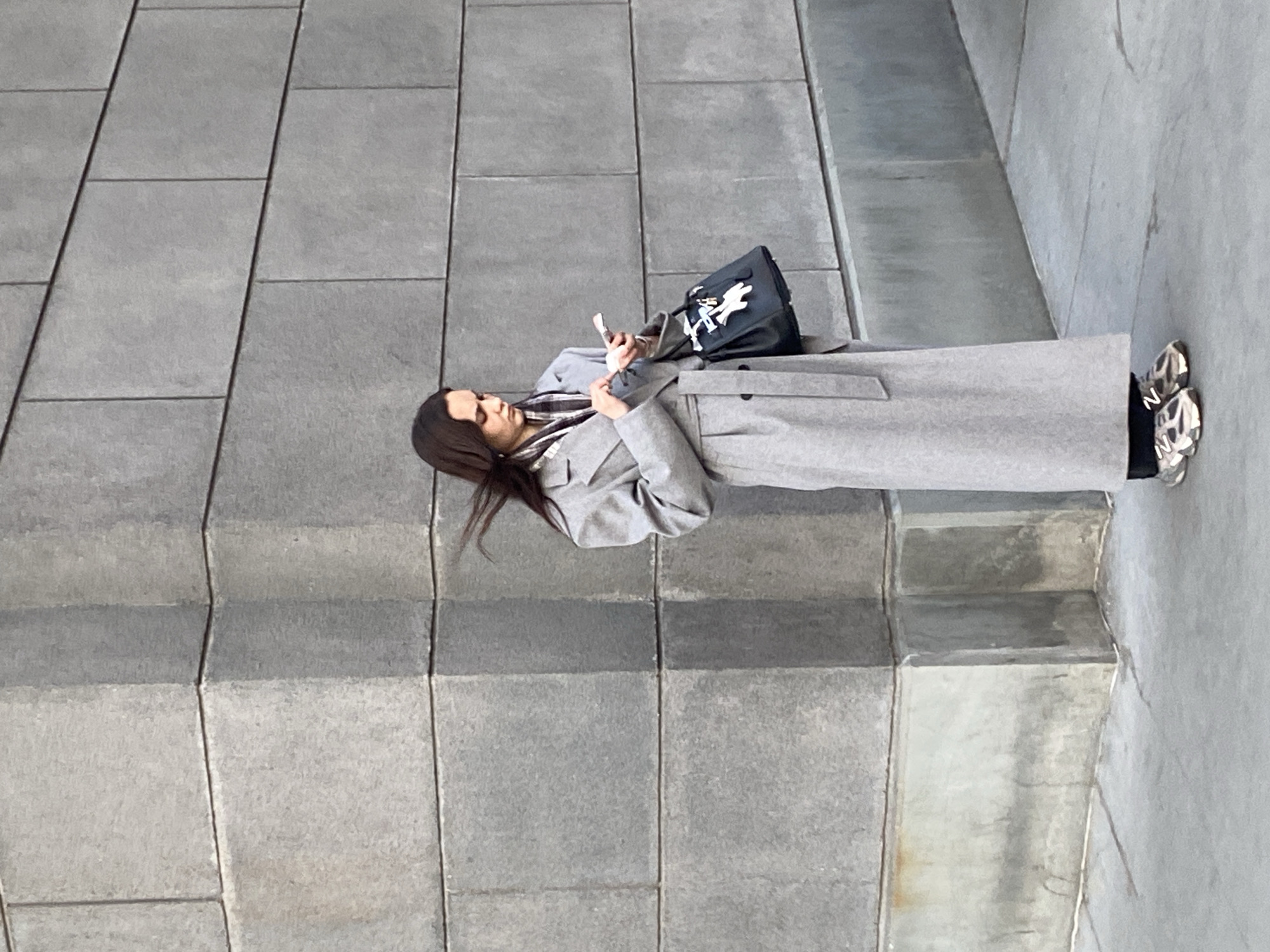 A female individual stands on a grey concrete sidewalk in Moscow, Russia, positioned against the corner of a building constructed from large, rectangular grey blocks. She has long, dark hair, which is ruffled by wind. She wears a full-length, light grey belted coat over a plaid scarf, dark trousers, and metallic-toned chunky athletic shoes. Both hands are occupied holding a white smartphone, with her gaze directed downwards at its screen. A black leather handbag, adorned with white motifs resembling a syringe, a stylized hand, and a small golden lock, is suspended from her left forearm. The lower section of the building's facade exhibits localized dark staining.