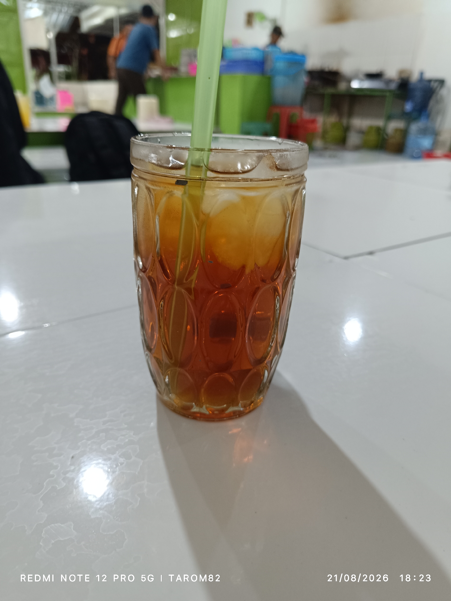 A close-up, eye-level shot shows a glass of iced tea on a white tiled surface. The glass is clear with a textured pattern and filled with a dark amber liquid and ice cubes. A light green straw extends from the glass. The background is blurred, showing what appears to be an indoor setting with people and various objects, possibly a food stall or a communal eating area in Sidodadi, Indonesia. The lighting is dim, with a bright reflection on the tiled surface in the foreground. The bottom right corner of the image displays a timestamp: "21/08/2026 18:23" and text indicating the device used: "REDMI NOTE 12 PRO 5G | TAROM82".