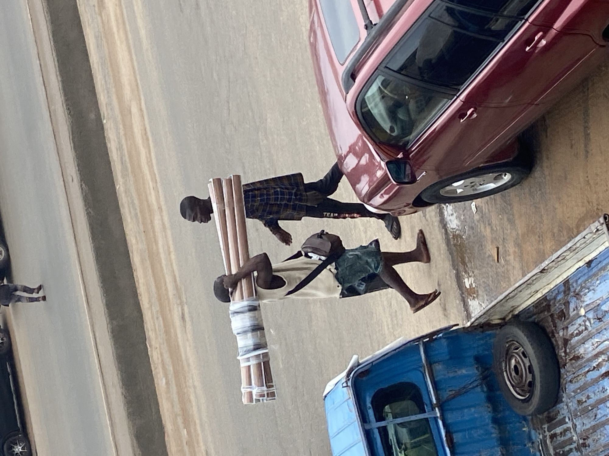 Two men are walking on a paved road in Ogbunike, Nigeria. The man on the left, wearing a sleeveless shirt and shorts, carries a bundle of long, cylindrical objects on his shoulders. He has a dark-colored bag slung across his chest. The man on the right, dressed in a plaid long-sleeved shirt and dark pants, walks beside him. A blue pickup truck is partially visible on the left side of the frame, with a spare tire in its bed. A red SUV is parked on the right side. The road appears wet in some areas, suggesting recent rain.
