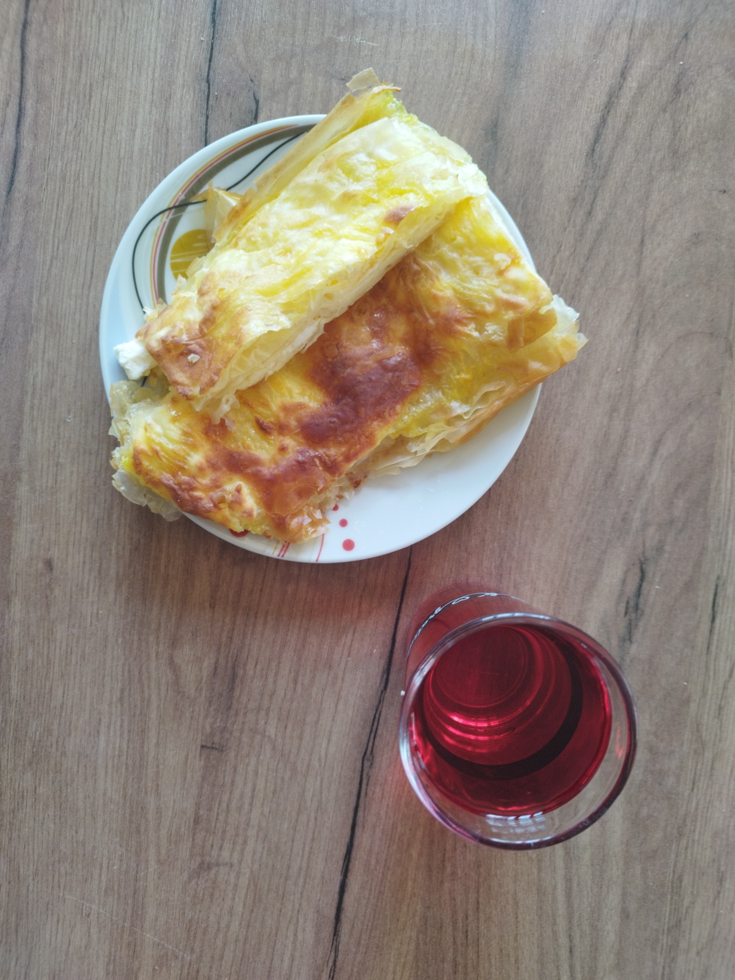 On a wooden surface, a plate of burek sits beside a glass of red liquid. The burek, a type of baked pastry, is golden and flaky, with visible layers. The plate has a white base with concentric circles of green, brown, and yellow around the edge, along with red dots. The glass, partially filled with a red beverage, is transparent, allowing the liquid’s color and the glass's shape to be clearly visible. The wooden surface has a light brown hue with a visible wood grain pattern.
There are no visible people in this scene. The setting appears to be indoors, possibly a dining table.
There is no information to indicate that this scene is in the city of Unknown, Unknown.