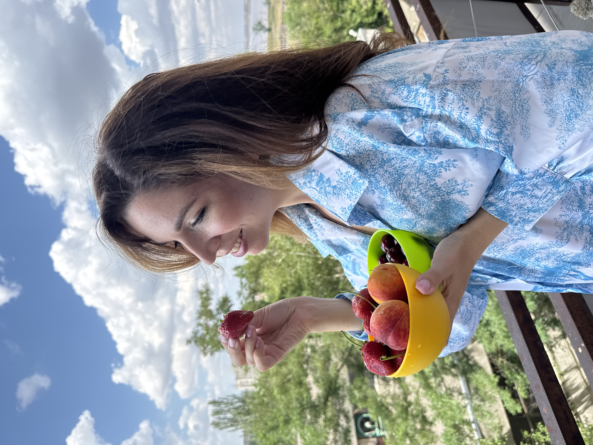 A woman stands on a balcony overlooking Odesa, Ukraine, holding a bowl of fruit. The woman, who has medium-length brown hair, is wearing a blue and white patterned shirt. She is holding a red strawberry in her right hand and a yellow bowl filled with strawberries and peaches in her left. Next to the yellow bowl, a green bowl contains cherries. The background shows trees, buildings, and a body of water under a cloudy blue sky.