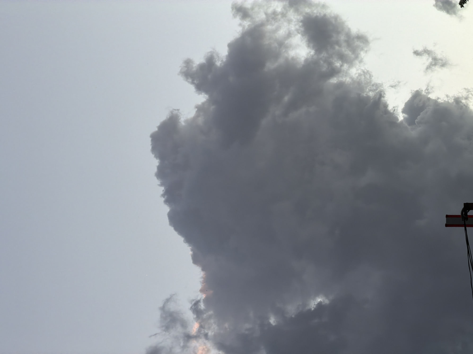 The image shows a cloudy sky, dominated by a large, dark cloud formation occupying the right side of the frame. The left side of the image shows a lighter, overcast sky. There is a partially visible object with a red stripe and cables on the right side of the image, possibly a piece of construction equipment or part of an urban structure. The location of the city of Sofia, Bulgaria is impossible to determine.