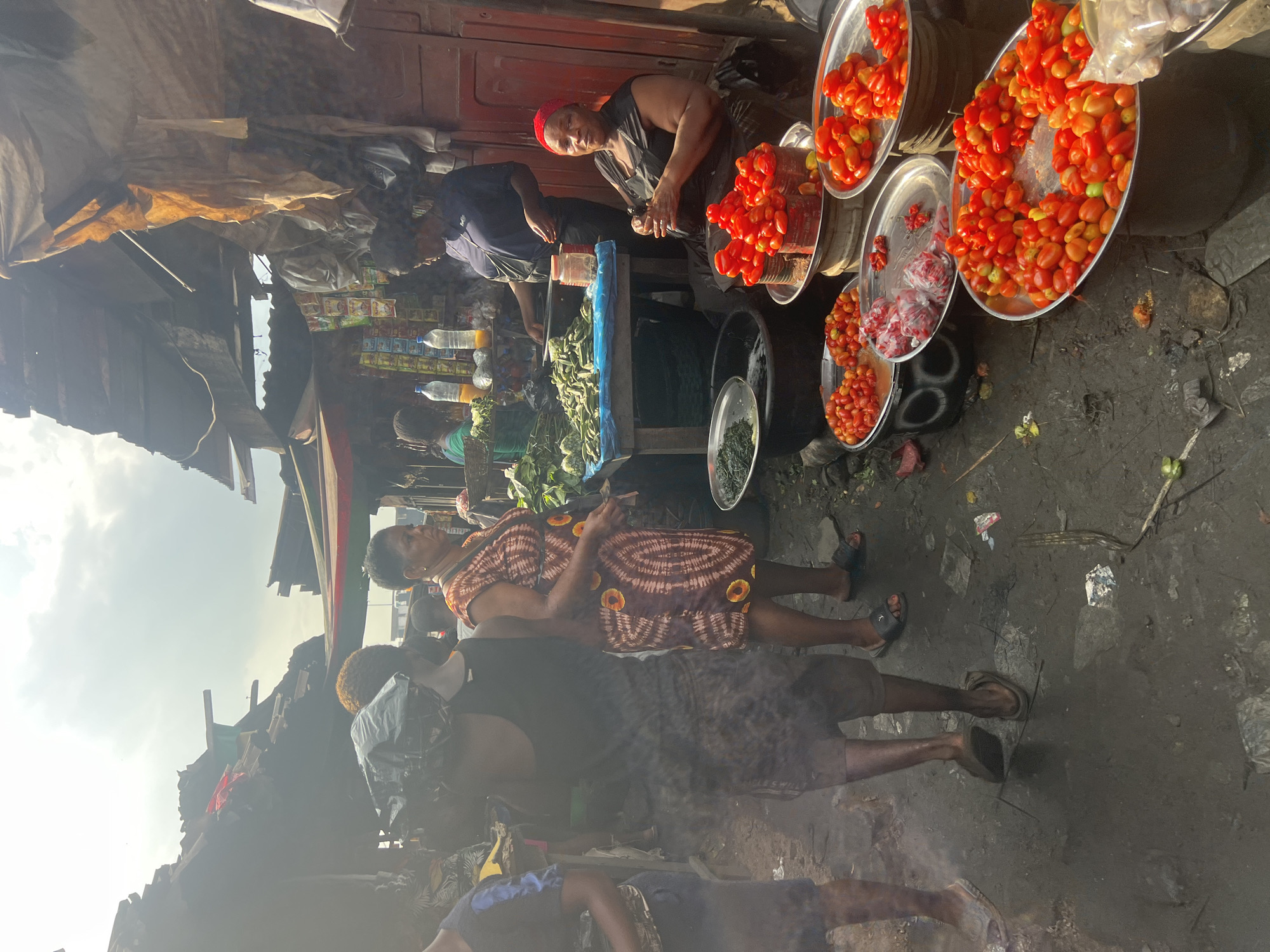 The image depicts a busy market scene in Onitsha, Nigeria. In the foreground, various fruits and vegetables are displayed in metal bowls, including tomatoes of different sizes and colors. A woman in a black sleeveless top and a red headscarf is seated, appearing to be selling produce. Behind her, another woman in a patterned dress stands, possibly interacting with a customer. In the background, more vendors and shoppers are visible, along with stalls laden with goods. The market is covered by makeshift roofs, and the ground is wet and muddy, suggesting recent rain. The overall atmosphere is one of lively commerce in an outdoor market setting.