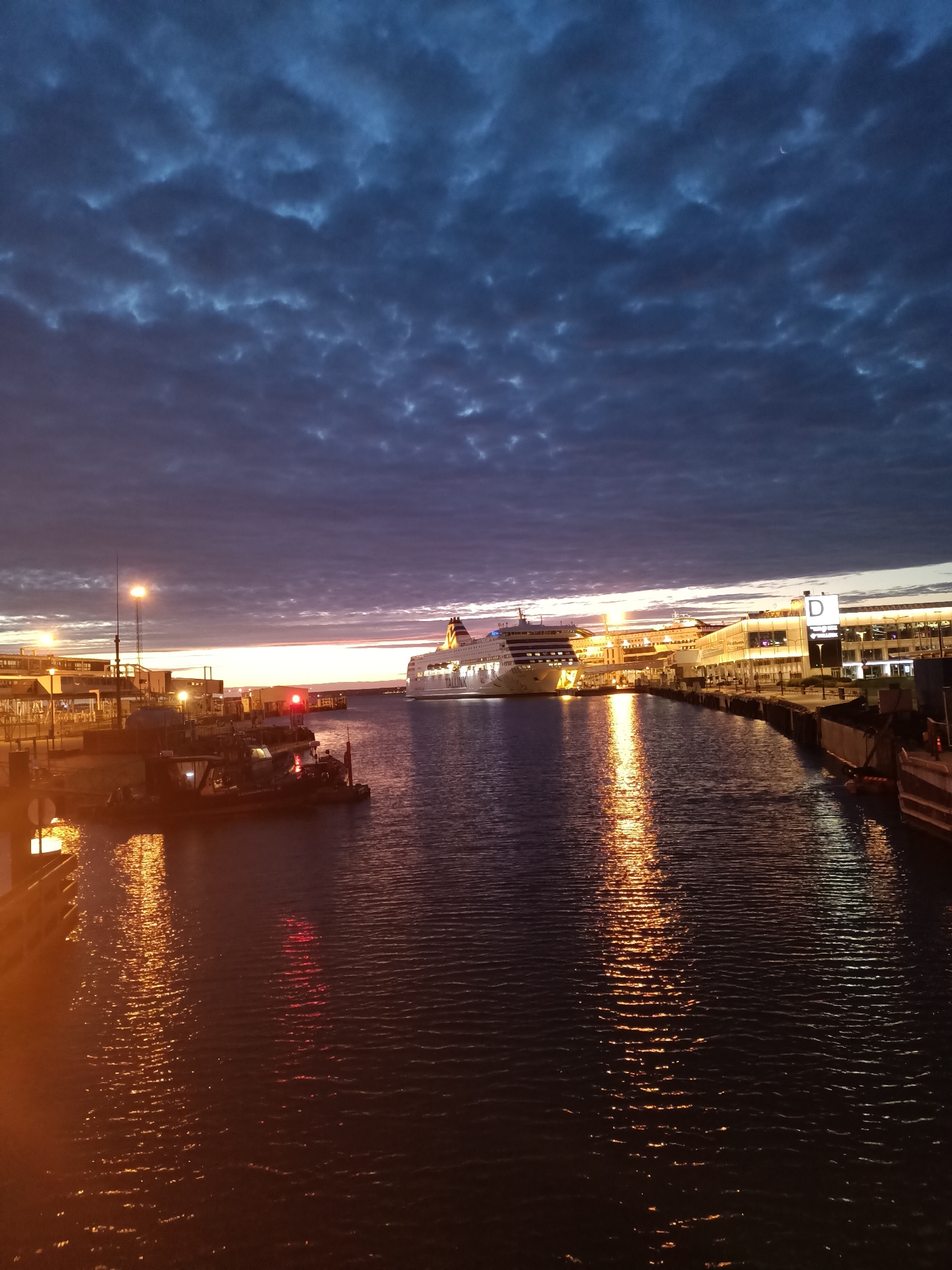 The image depicts the Tallinn Passenger Port in Estonia at dusk. A large cruise ship, likely a ferry, is docked or approaching a terminal on the right side of the frame. The terminal building is illuminated, with the letter "D" prominently displayed on a sign. Several smaller vessels, possibly tugboats or service boats, are moored in the foreground. The sky is filled with dramatic, dark clouds, with streaks of orange and pink light indicating the setting sun near the horizon. The water reflects the lights from the ships and the terminal, creating shimmering patterns. The overall atmosphere is serene and maritime, characteristic of a port at twilight.