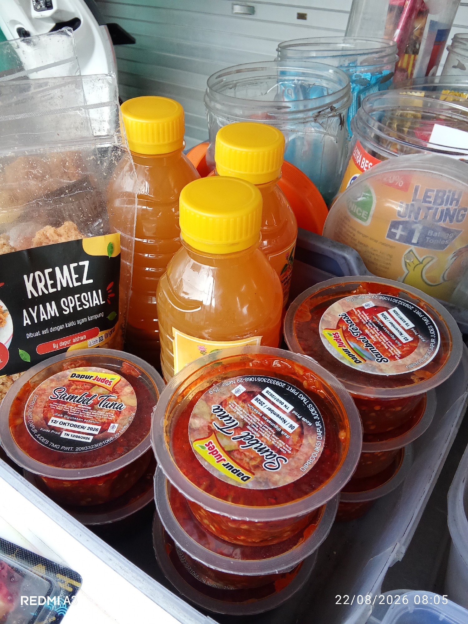 The image captures a close-up, slightly angled view of several food products arranged on a surface, likely a market stall or countertop. In the foreground, there are multiple round, clear plastic containers filled with a reddish-brown condiment, which appears to be sambal. The labels on these containers indicate "Sambal Tuna" and "Dapur Judes." To the left, a clear plastic bag contains what looks like crispy fried chicken pieces, with a black label that reads "KREMEZ AYAM SPESIAL." Behind these, there are several glass bottles with yellow caps, containing an amber-colored liquid, possibly a sauce or drink. In the background, various glass jars, some empty and others containing items or labels, are visible. One label clearly shows "LEBIH UNTUNG +1 Bantal Toples" with a cartoon illustration. The overall setting suggests a food retail environment. The timestamp "22/08/2026 08:05" is present in the bottom right corner, and "REDMI A3" is visible in the bottom left, indicating the device used for capture. The scene does not show any people or specific city landmarks to confirm its location in Cemani, Indonesia, beyond what might be inferred from product labeling or context not provided in the image.