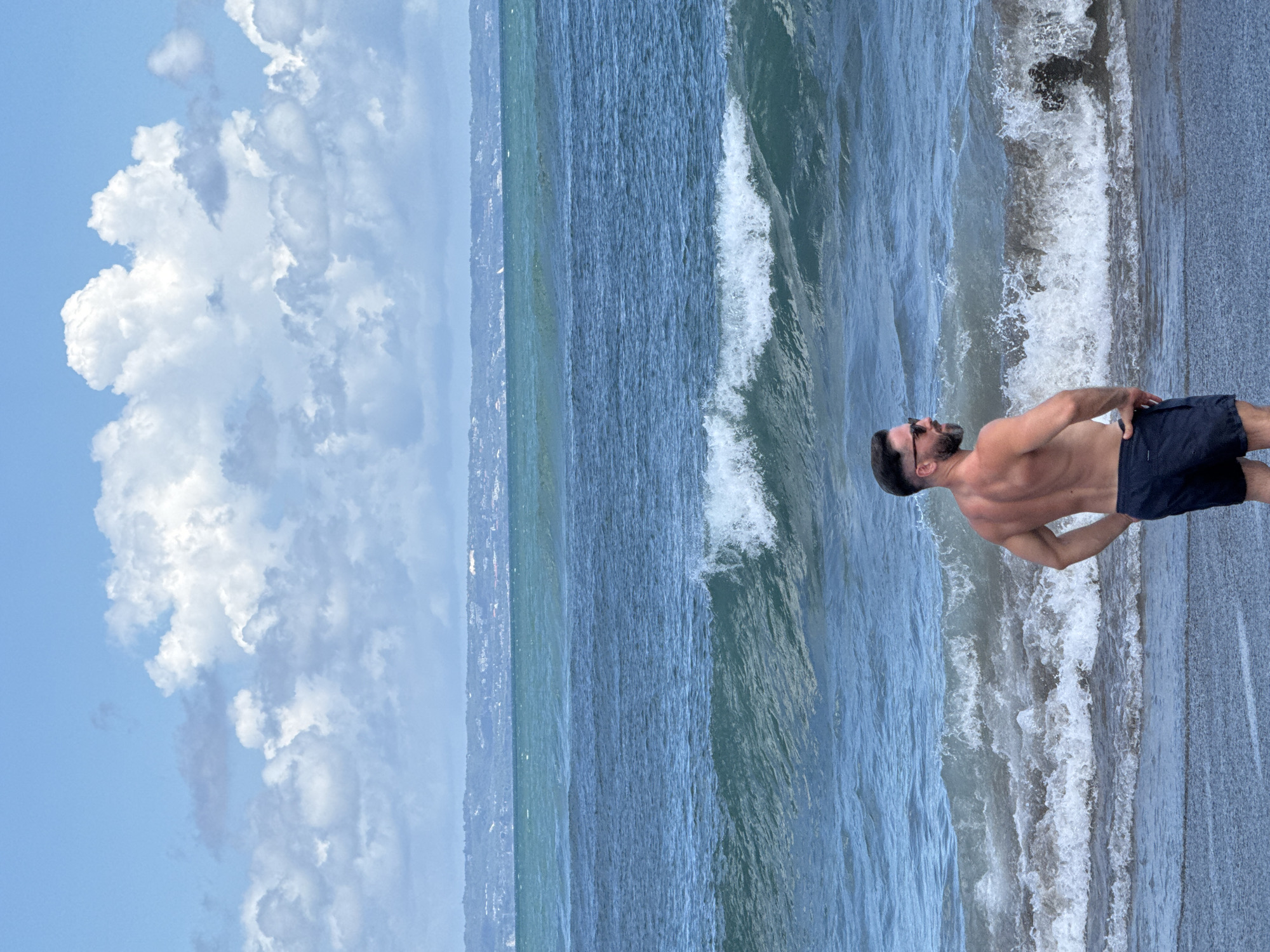 A shirtless man in dark swim trunks stands on a beach with his hand on his hip, seemingly gazing out at the ocean. Behind him, waves are crashing onto the shore. The sky is partly cloudy, with puffy white clouds occupying the left side of the frame. In the distance, a strip of land is visible along the horizon. This appears to be a scene along a beach in North Kuta, Indonesia.