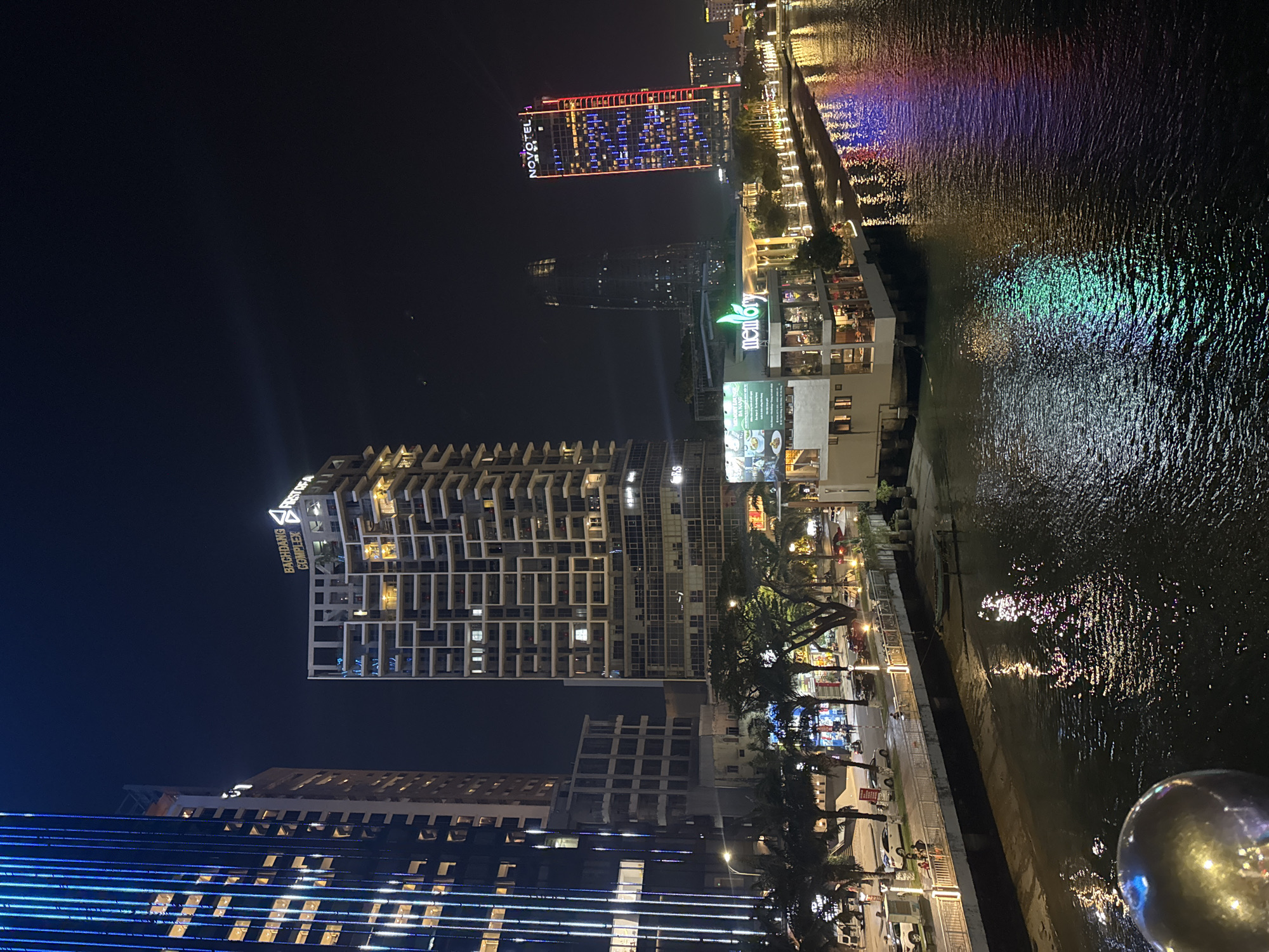 At nighttime in Đà Nẵng, Vietnam, the illuminated Novotel hotel is visible next to the Han River. The river's surface reflects the lights from the city, including colorful reflections. The Bach Dang Complex building is located nearby, as well as the Memory restaurant. There are palm trees along the riverside, and lights illuminate the street.