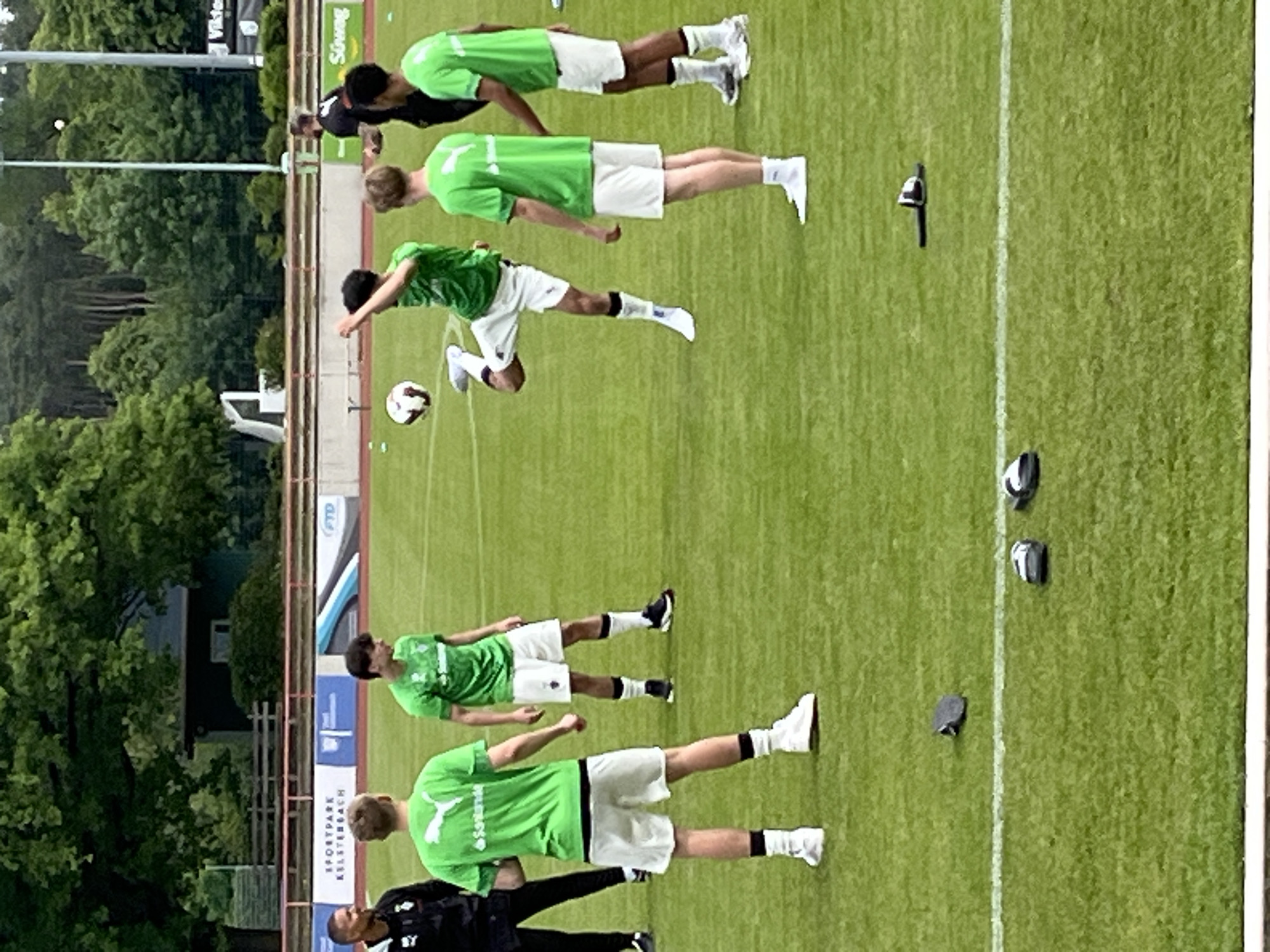 The image shows a soccer training session at Sportpark Kelsterbach in Kelsterbach, Germany. There are six soccer players, five wearing green shirts and white shorts, and one wearing a black tracksuit. One player is kicking a soccer ball in the air. The training session is taking place on a green soccer field next to a white line. There are several black objects near the white line. There are trees and buildings in the background.