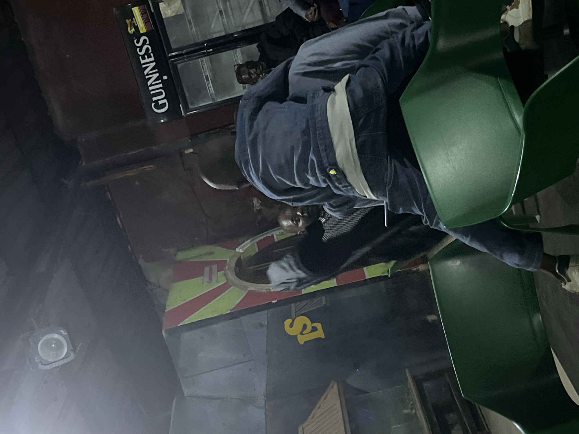 The image depicts an indoor scene at what appears to be a bar or lounge in Jos, Nigeria. In the foreground, a young child wearing a blue denim jacket and pants is seated on the lap of an adult, whose legs are also visible. The child is facing away from the camera, towards a man wearing glasses and a dark jacket. To the left, a large wall decoration with red and green diagonal stripes is visible, with a yellow "LS" logo. Behind the man, a Guinness branded refrigerator is present, indicating a place that sells beverages. Other individuals are dimly visible in the background, and the lighting suggests it is nighttime or a dimly lit interior. The environment seems somewhat cluttered, with green plastic chairs in the foreground and other furniture or structures in the background.