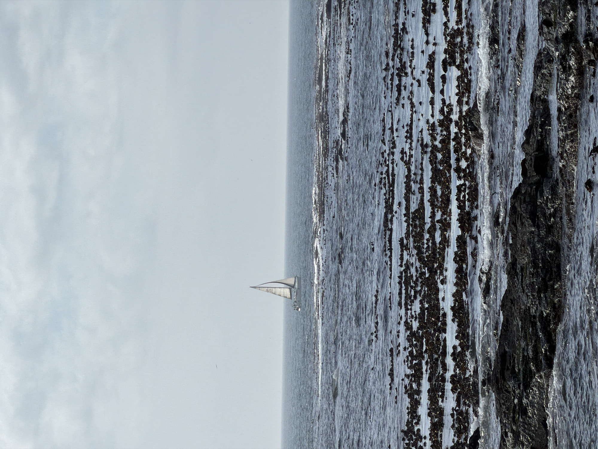The image depicts a seascape, with a sailboat visible on the horizon. The right side of the frame shows a rocky coastline. The left side features a cloudy sky. The scene is oriented vertically. The location appears to be Cape Town, South Africa, given the coastline.