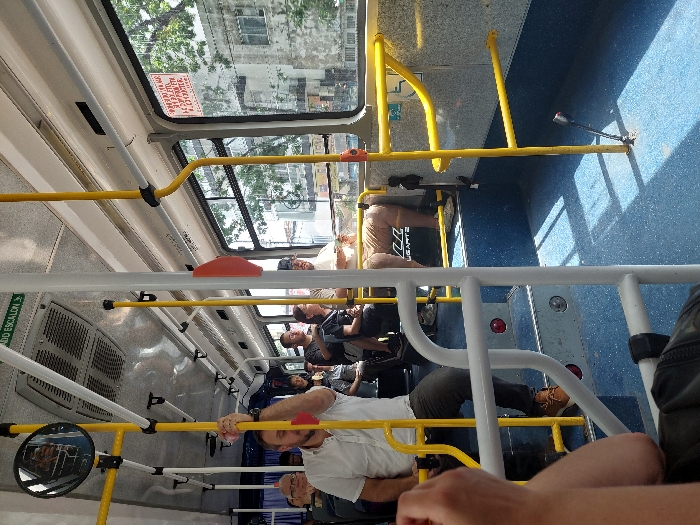 The image captures the interior of a public transit bus on a bright, sunny day, looking from the rear towards the front. Several passengers are visible, either seated or standing and holding onto the prominent yellow grab bars and white handrails that crisscross the cabin.
In the foreground, a man in a white t-shirt and dark pants is standing, holding a yellow bar. His legs and feet, wearing brown boots, are visible near the bottom right. Further inside the bus, other passengers are scattered. One man in a light brown shirt is seated facing away from the camera, while another in a dark shirt is seated further back. The blue-patterned floor is brightly illuminated by sunlight streaming through the windows, casting distinct shadows of the bus's structure and passengers.
Through the large windows, an urban environment can be seen, with green trees and buildings under a clear sky. The bright conditions suggest it is daytime, likely morning or afternoon.
Notable details include a red warning sign above a window that reads "ATENCION ALTO IMPACTO EN LA CABEZA" (Attention high impact to the head), indicating a potential hazard. On the left side, a green sign with partially visible text reads "CUIDADO ESCALON" (Watch your step). A small, circular mirror is mounted on a yellow pole to the left, reflecting a portion of the bus interior.
The overall scene depicts a typical commute or travel moment within a bus in an urban area, consistent with the provided location context of Buenos Aires, Argentina. Passengers appear neutral, engaged in their journey.
