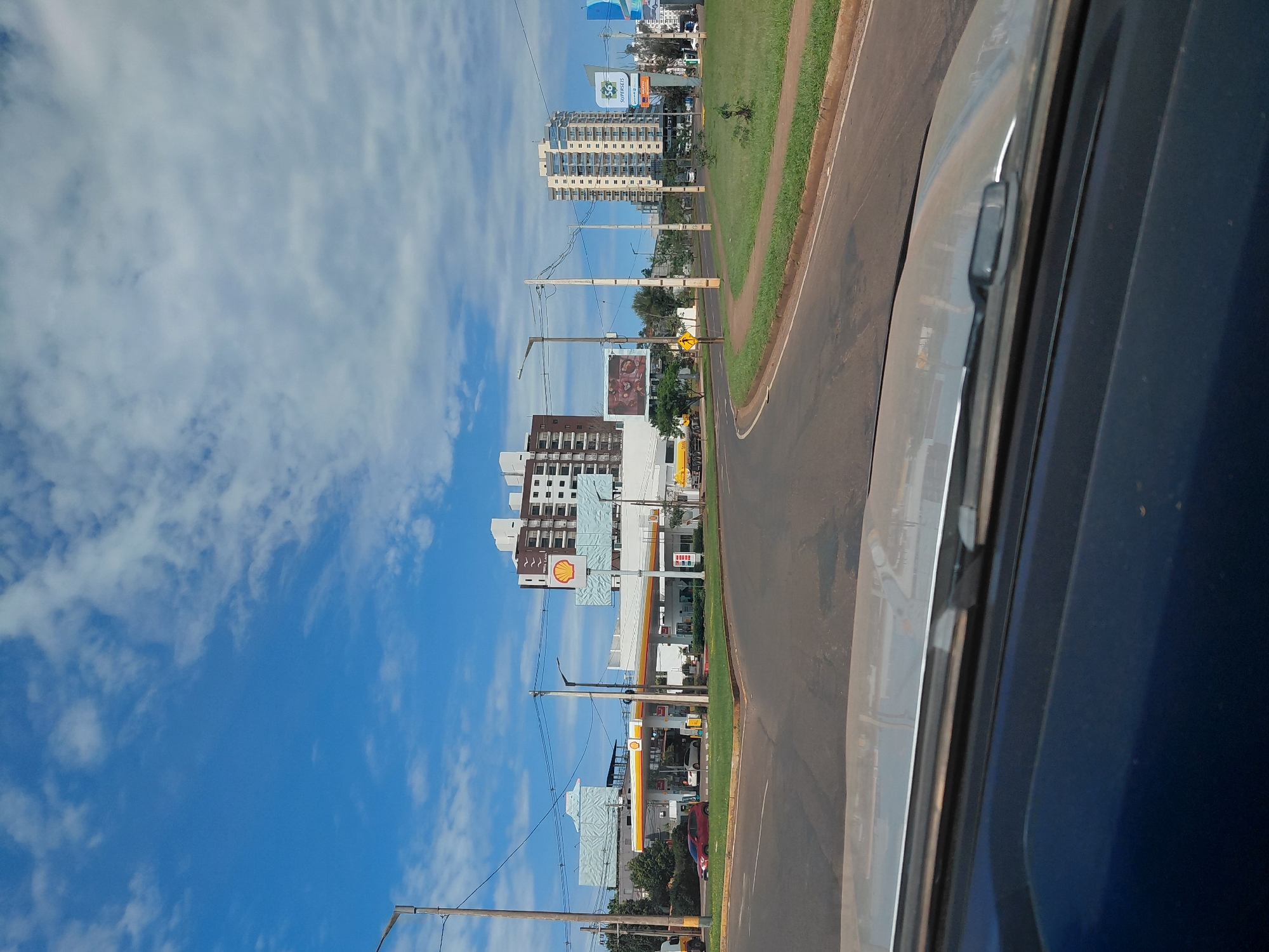 The image captures a street scene in Encarnacion, Paraguay, on a bright, cloudy day. In the foreground, the dashboard and windshield of a vehicle are visible, suggesting the photo was taken from inside a car. The road curves to the right, bordered by a green grassy median.
To the left of the road, a Shell gas station is prominent, with its distinctive yellow and red canopy and signage. Several cars are parked at the pumps. Beyond the gas station, a multi-story building with a patterned facade stands, and further back, two taller, modern apartment buildings are visible.
On the right side of the road, another tall apartment building with balconies can be seen, along with billboards and signage for businesses. A yellow truck is also visible in the distance on the right. The sky is a vibrant blue, dotted with white, fluffy clouds. The overall impression is one of an urban or suburban commercial area on a clear day.