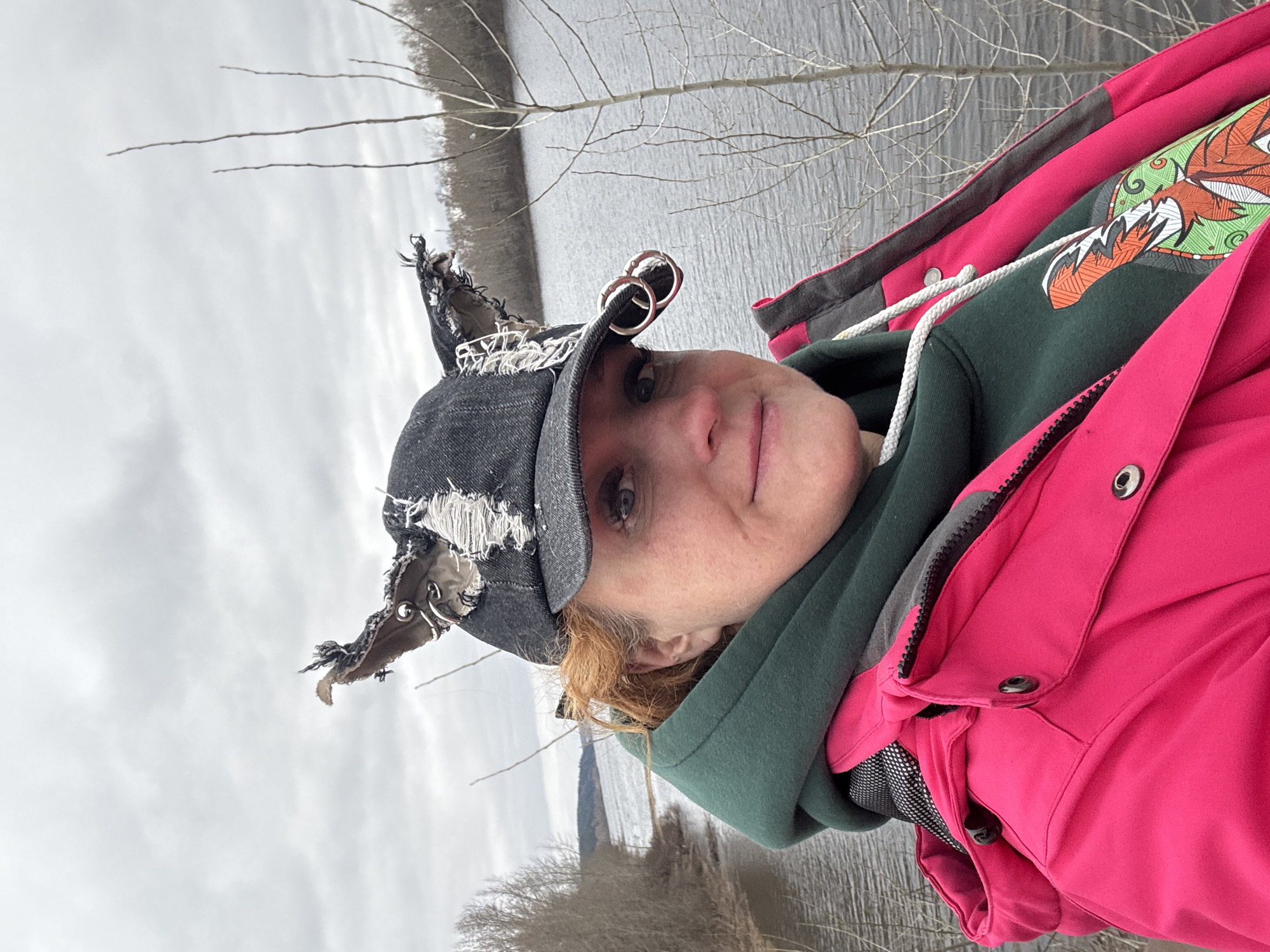 A woman is pictured outdoors against a river backdrop. She is wearing a black, distressed baseball cap with silver hoops attached to the bill, a green hooded sweatshirt featuring an image of a fox, and a bright pink jacket. The environment includes a body of water, bare trees, and an overcast sky. The location is Chaikovsky, Russia.