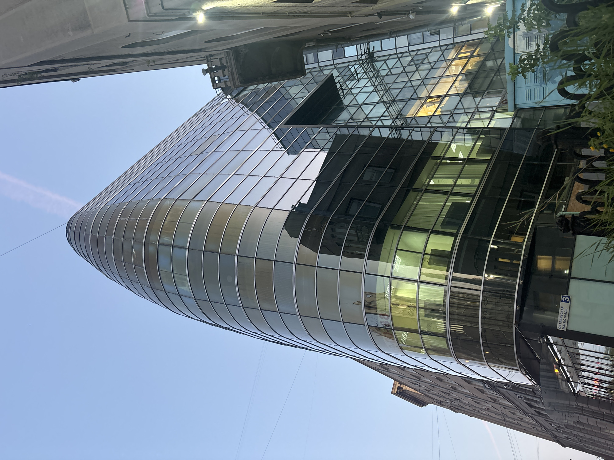The image shows a modern glass-fronted building juxtaposed with an older, stone building. The glass building has a curved facade that reflects the sky and the adjacent building. The older building has a more traditional architectural style with visible windows and balconies. The scene is captured from a low angle, looking upwards towards the buildings. A street sign that reads "ОКТЯБРЬСКАЯ МАГИСТРАЛЬ 3" is visible on the glass building, indicating the location as Oktyabrskaya Magistral, house number 3. The sky is clear and blue, suggesting it is daytime. The overall impression is an urban environment with a mix of old and new architecture.