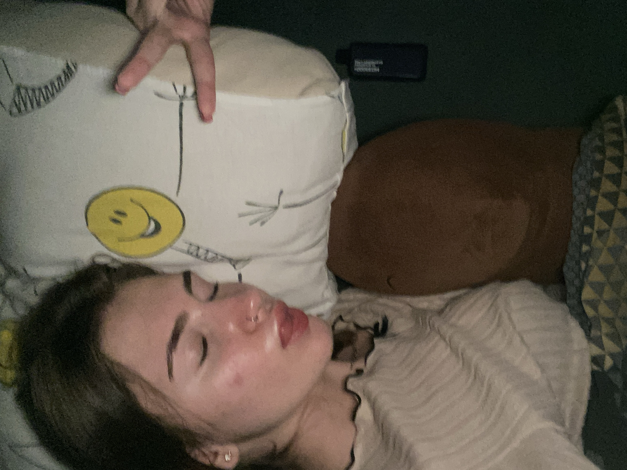 A young woman with dark hair and closed eyes is lying on a pillow with a smiley face on it. Her head is tilted to the side, and her lips are slightly parted. She is wearing a light-colored, ruffled shirt. A hand with painted nails is making a peace sign in the background, positioned over the pillow. A dark bottle is visible on the dark green surface next to the pillow. The scene appears to be indoors, possibly a bedroom.