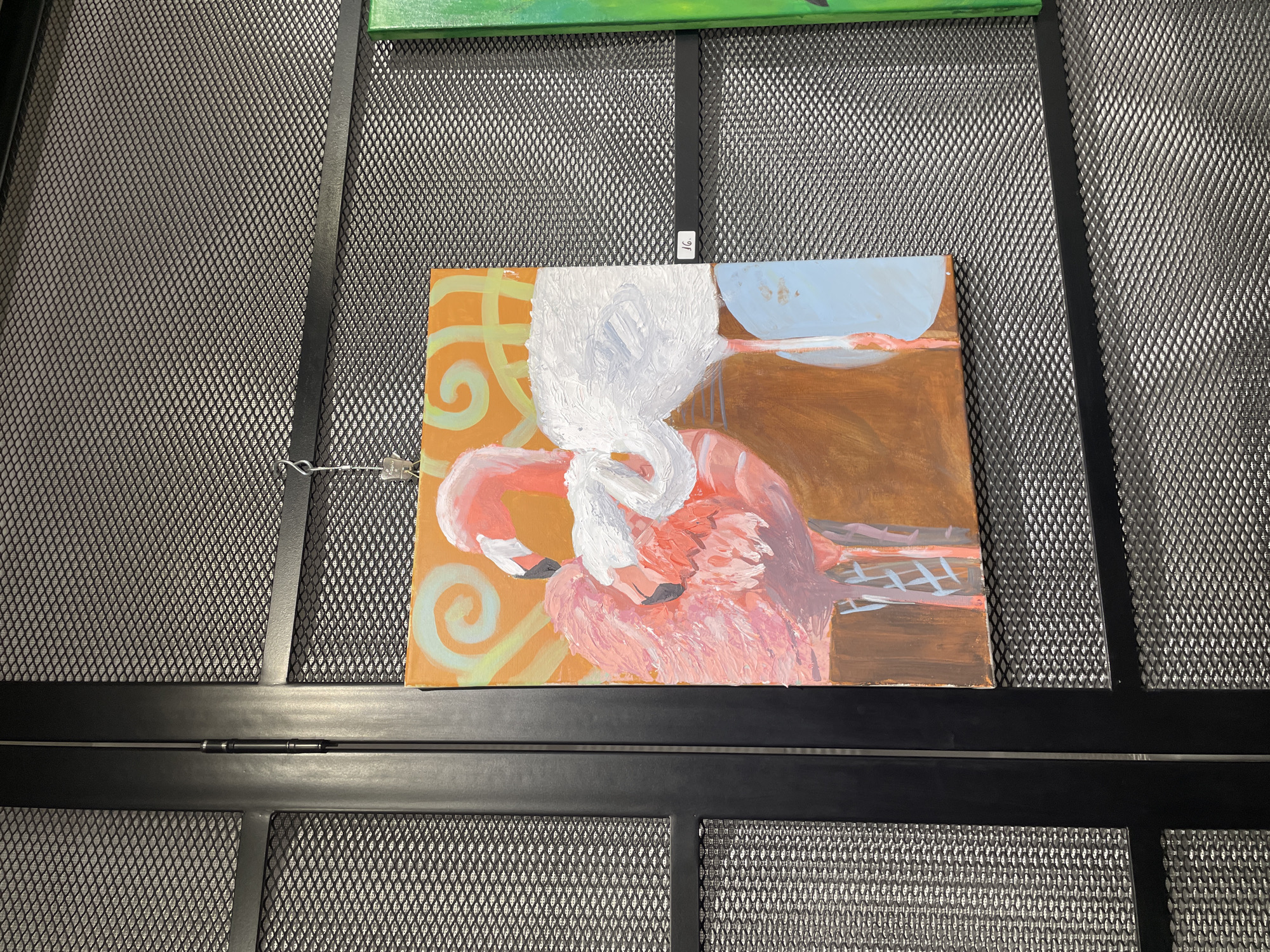 The image shows a painting of two flamingos hanging on a black metal mesh display in Plovdiv, Bulgaria. The painting is square and has an orange background with yellow swirls on the left side. The flamingos are rendered with broad brushstrokes, predominantly in shades of pink, white, and orange. The background transitions to brown with a blue area suggesting the sky or water. A small white tag is attached to the top of the painting, indicating it is for sale. The mesh display is part of an art exhibition.