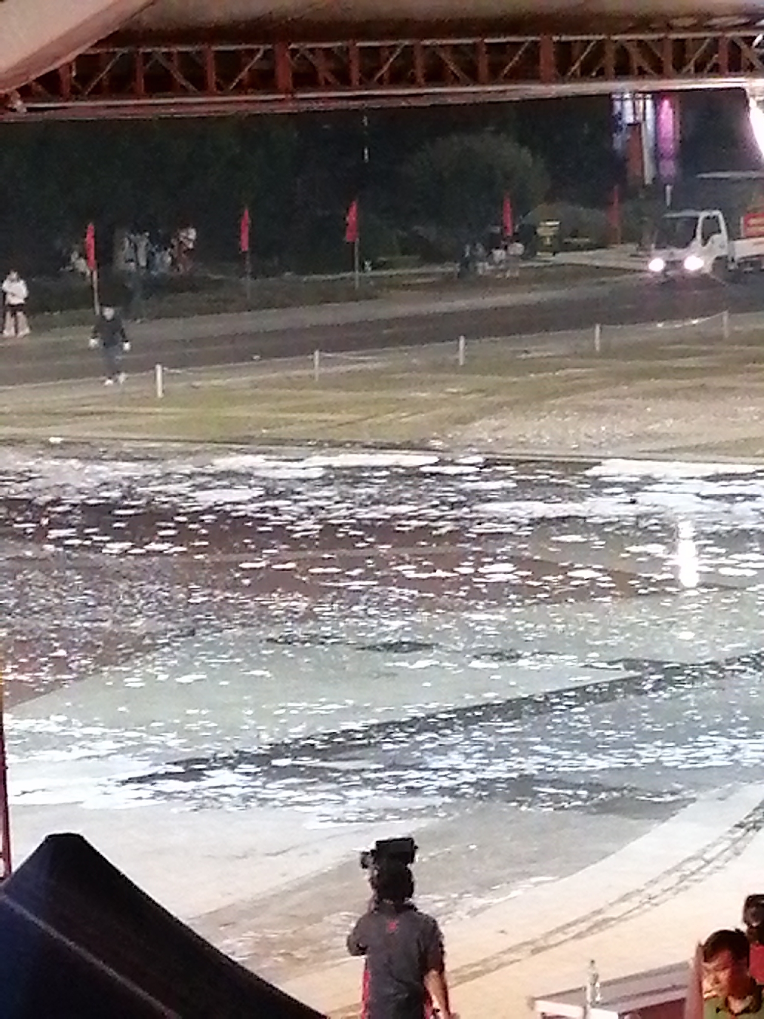 In Da Lat, Vietnam, an outdoor event is underway, possibly a performance or a parade, under a covered structure with exposed metal beams. The scene features a large, reflective surface, perhaps a flooded area or a temporary water feature, covering the foreground. White, irregular shapes are scattered across this surface, creating a textured appearance.
In the background, a group of people are gathered, some standing and some walking. Red flags are visible, planted in the ground, suggesting a demarcated area or part of the event's decoration. Further back, a white truck with its headlights on is present, indicating it might be daytime or early evening, with artificial lighting being used. The ground beyond the reflective surface appears to be grassy or dirt.
In the foreground, a person is seen from behind, wearing dark clothing and a camera on their head, likely documenting the event. Another person, also in dark clothing, is partially visible in the lower right corner. The overall atmosphere appears to be lively, with an event in progress, though specific details about the nature of the event are not clear. The time of day is difficult to determine precisely, but the truck's headlights suggest it's not the middle of the day. The weather is not explicitly discernible from the image.