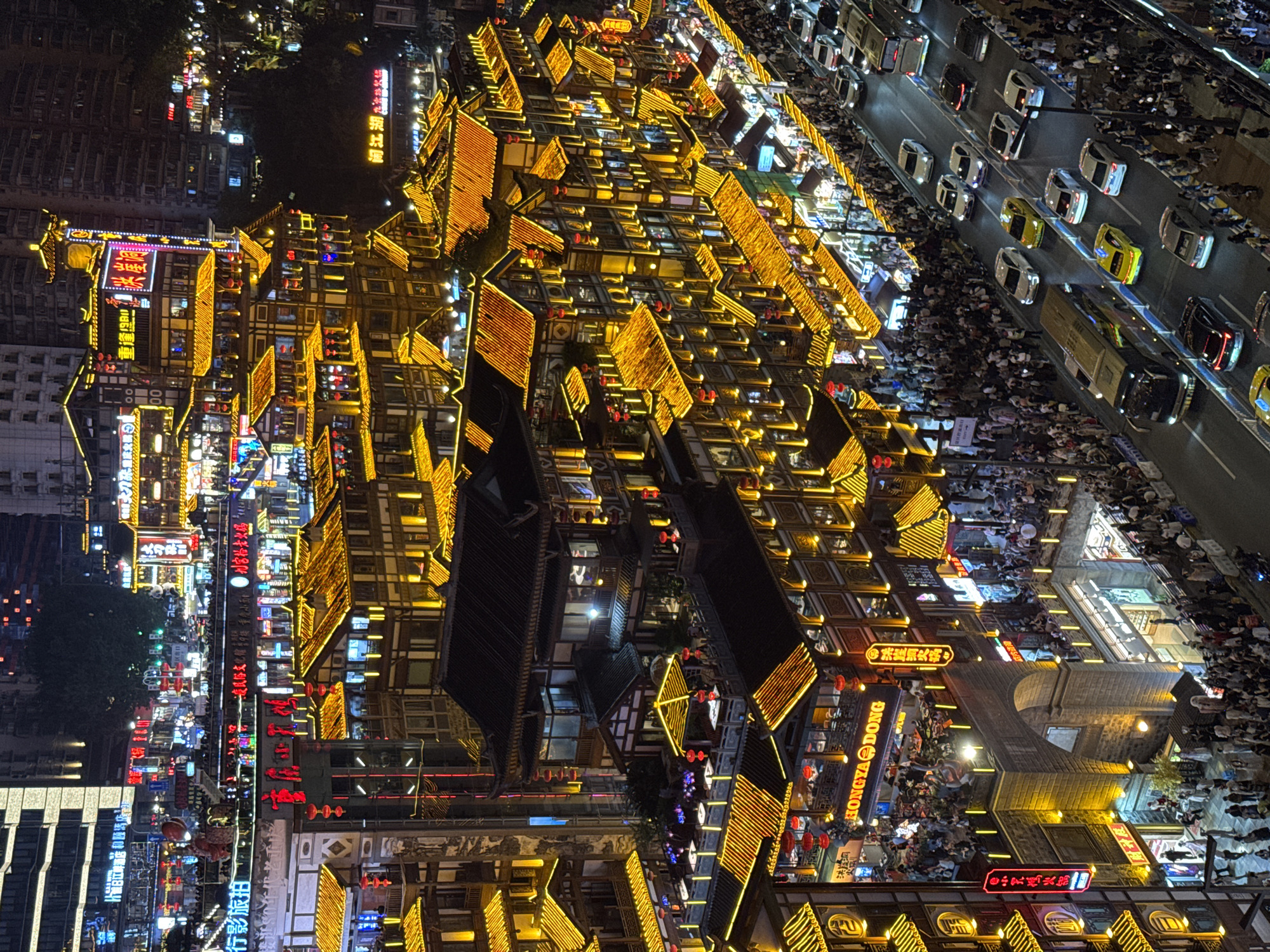 This nighttime aerial photograph depicts the illuminated cityscape of the Hongyadong commercial and residential complex located in the Yuzhong District of Chongqing, China. The buildings are distinguished by traditional Chinese architectural styles. The structure is accented by golden lights outlining the edges of the roofs and windows, a road parallels the district with cars and crowds of people are visible along the side of the road and between the buildings. Red Chinese lanterns can be seen interspersed among the buildings. In the lower left, commercial signs are illuminated in various colors, including red, white, and blue. The overall ambiance is lively and vibrant.