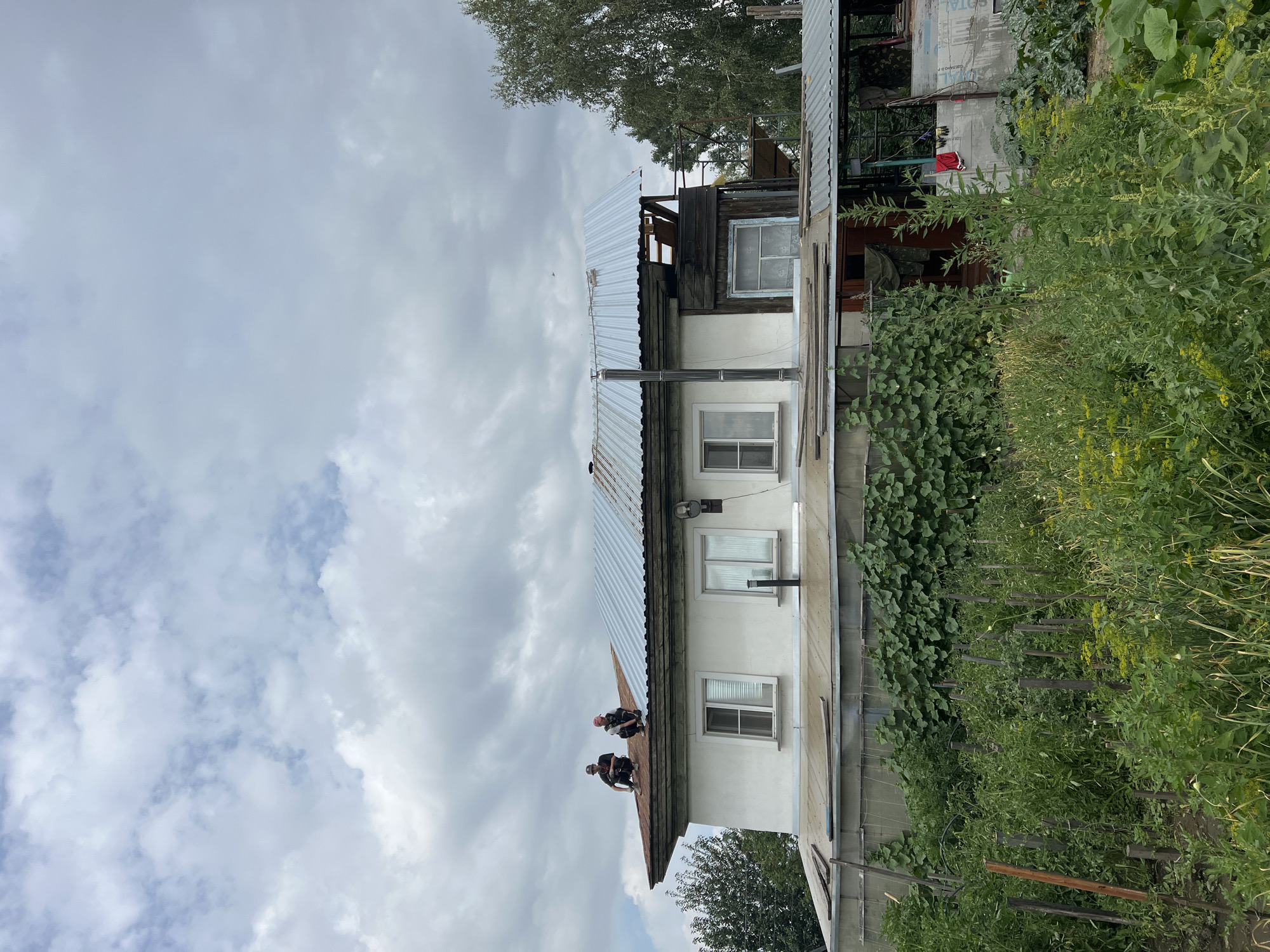 Two men are working on the roof of a two-story house in Biysk, Russia. The house has a white facade and a white corrugated metal roof that is partially installed. The lower section of the house is covered by a green greenhouse structure. In front of the house is a garden with various plants and wooden stakes. The sky is cloudy.