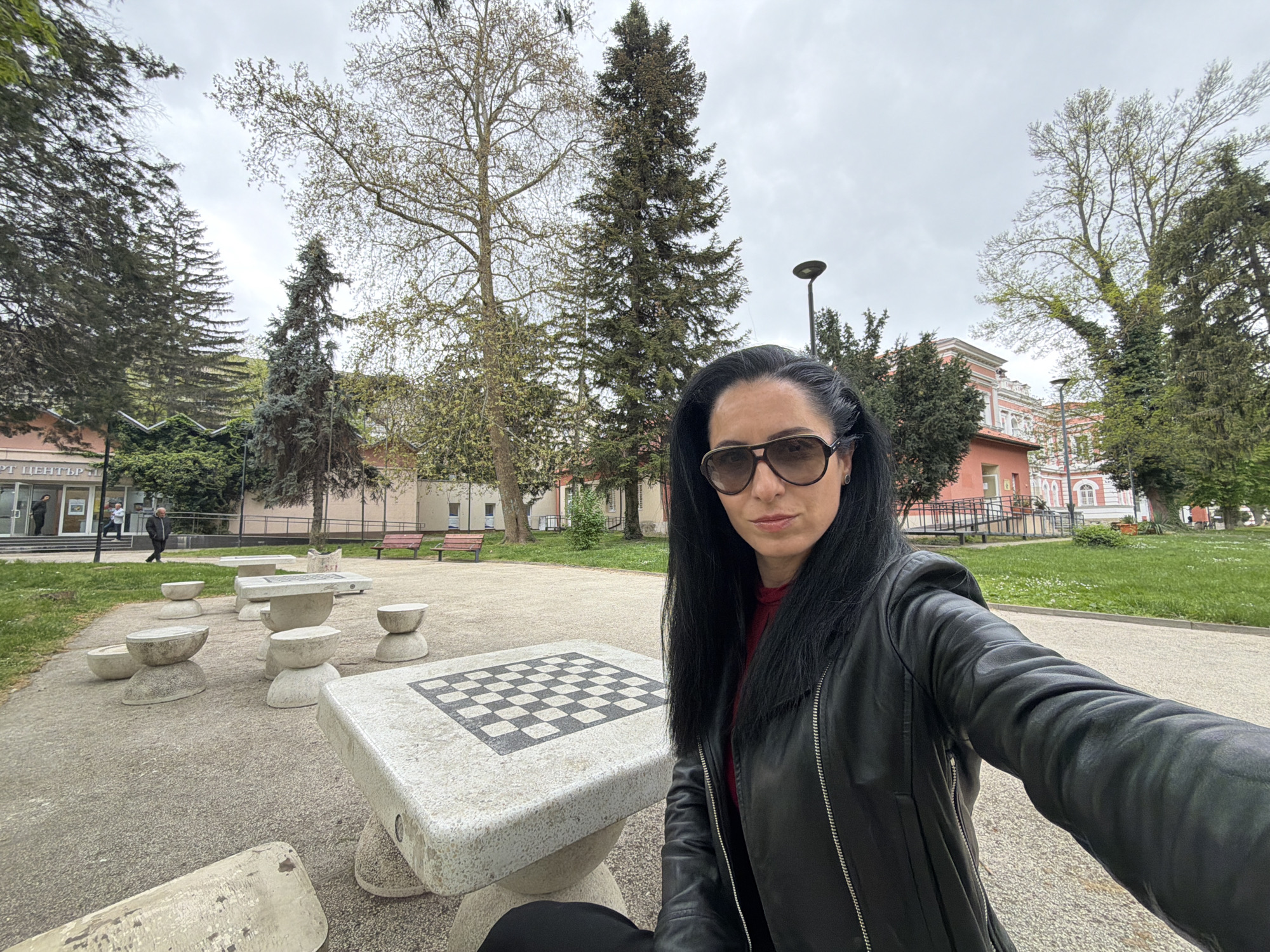 The image captures a selfie of a woman in a public park or square in Pleven, Bulgaria, on an overcast day. The main subject, a woman with long, straight dark hair, wears dark sunglasses and a black leather jacket over a red top, holding the camera for a selfie with a neutral expression.
The scene features a paved outdoor area with several stone tables and seats. A large stone table with a black and white chessboard pattern is prominent in the foreground, with unique stone seats shaped like stacked hemispheres nearby. Beyond this paved section, there's a grassy lawn dotted with various trees, including tall, dark conifers and deciduous trees showing new, light-green leaf growth, indicating spring. Streetlights and several park benches are also visible.
In the background on the left, a light-colored building with steps leading to an entrance is seen, near which two men are present – one walking, another near the steps. Further to the right, a distinctive older building with pink-red walls, white architectural details, and a ramp leading up to it adds to the background.
The sky is cloudy and grey, suggesting an overcast day without direct sunlight. On the building in the left background, partial Cyrillic text is visible, reading "РТ ЦЕНТЪР", likely part of a larger sign such as "СПОРТЕН ЦЕНТЪР" (Sports Center).