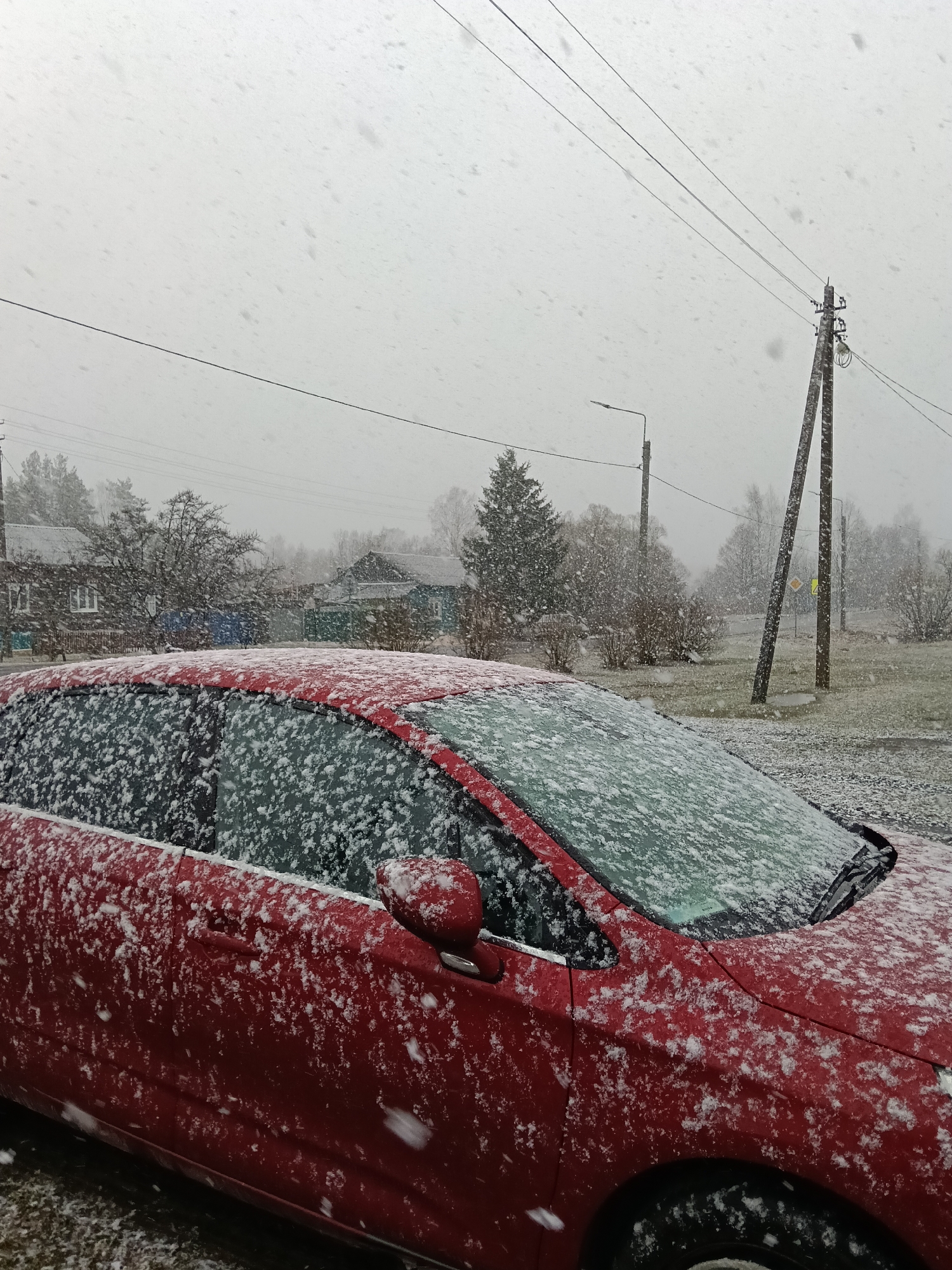 The image shows a red car covered in snow. The car is parked on a street in Красный Маяк, Russia. In the background, there are wooden houses, trees, and snow-covered ground. Power lines and utility poles are also visible. It is snowing heavily. No people are visible.