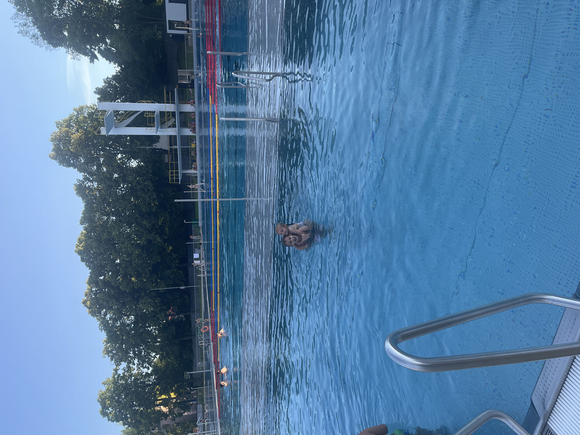 In a public swimming pool, a young boy gives a piggyback ride to a younger girl in the foreground. The water is a vibrant blue, reflecting the clear, sunny sky and the surrounding green trees. In the background, a diving platform stands tall, with swimmers visible in different lanes of the pool, marked by red, yellow, and blue buoys. The scene evokes a sense of a typical summer day at a recreational facility.