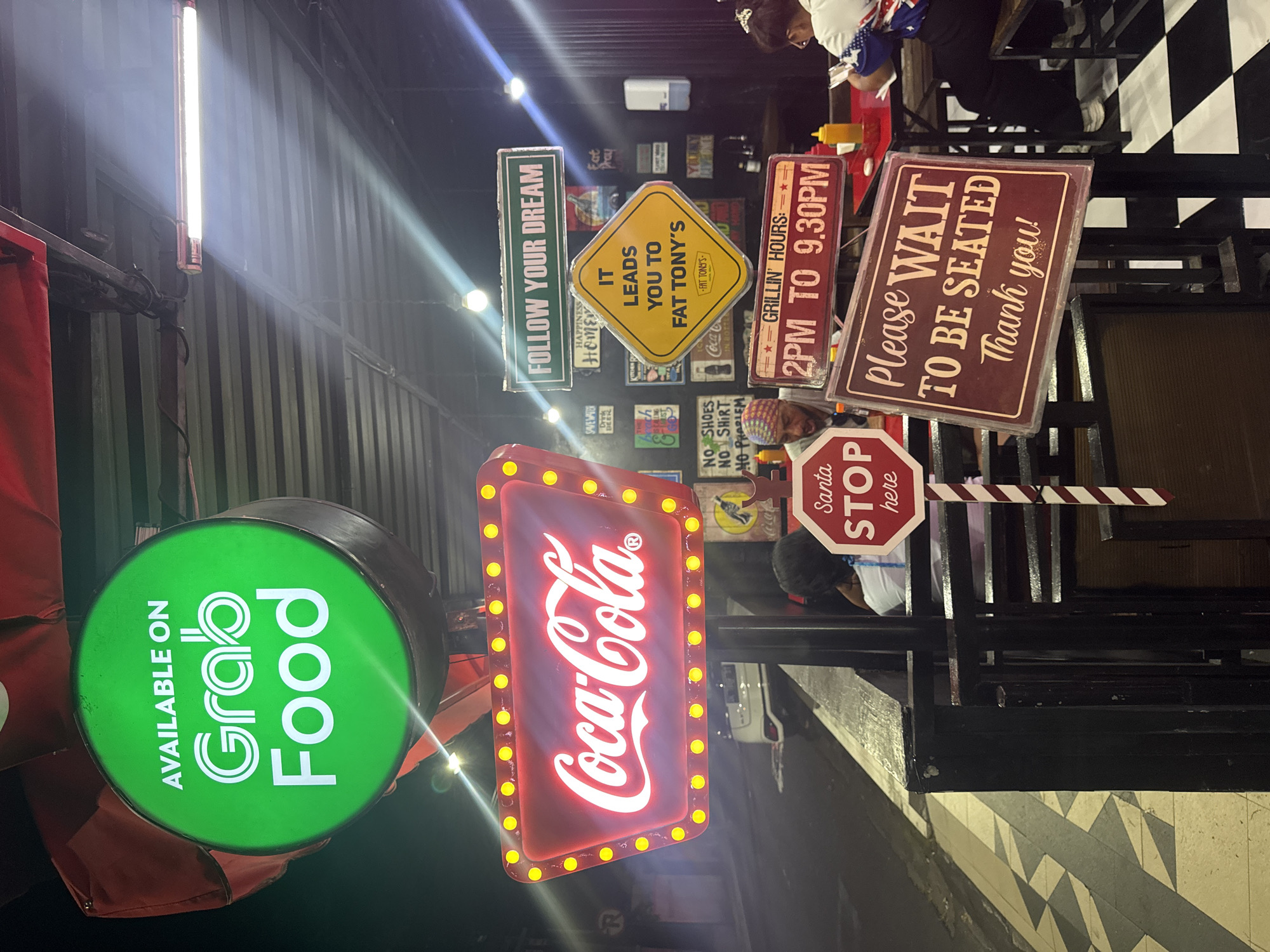 This photo captures a vibrant, brightly lit exterior of a food establishment, possibly a restaurant or eatery, likely located in Kuta, Indonesia. Several eye-catching signs vie for attention. At the forefront is a circular green sign reading "Available on Grab Food," indicating that the establishment offers delivery service through the Grab platform. Adjacent to it, a red Coca-Cola sign with a border of illuminated yellow lights adds a classic, nostalgic touch. Various smaller signs decorate the wall, including "Follow Your Dream," "It Leads You To Fat Tony's," "Grillin' Hours: 2PM to 9:30 PM," and "Please Wait To Be Seated Thank You!". A Santa "Stop Here" sign is also visible.
The environment appears to be an outdoor or semi-outdoor seating area with a checkered floor pattern. Parts of people seated at the tables are visible. A fluorescent light fixture on the wall provides additional illumination. The composition of the photo provides a close, detailed view of the signage, while the presence of customers hints at the active, bustling atmosphere of the establishment.