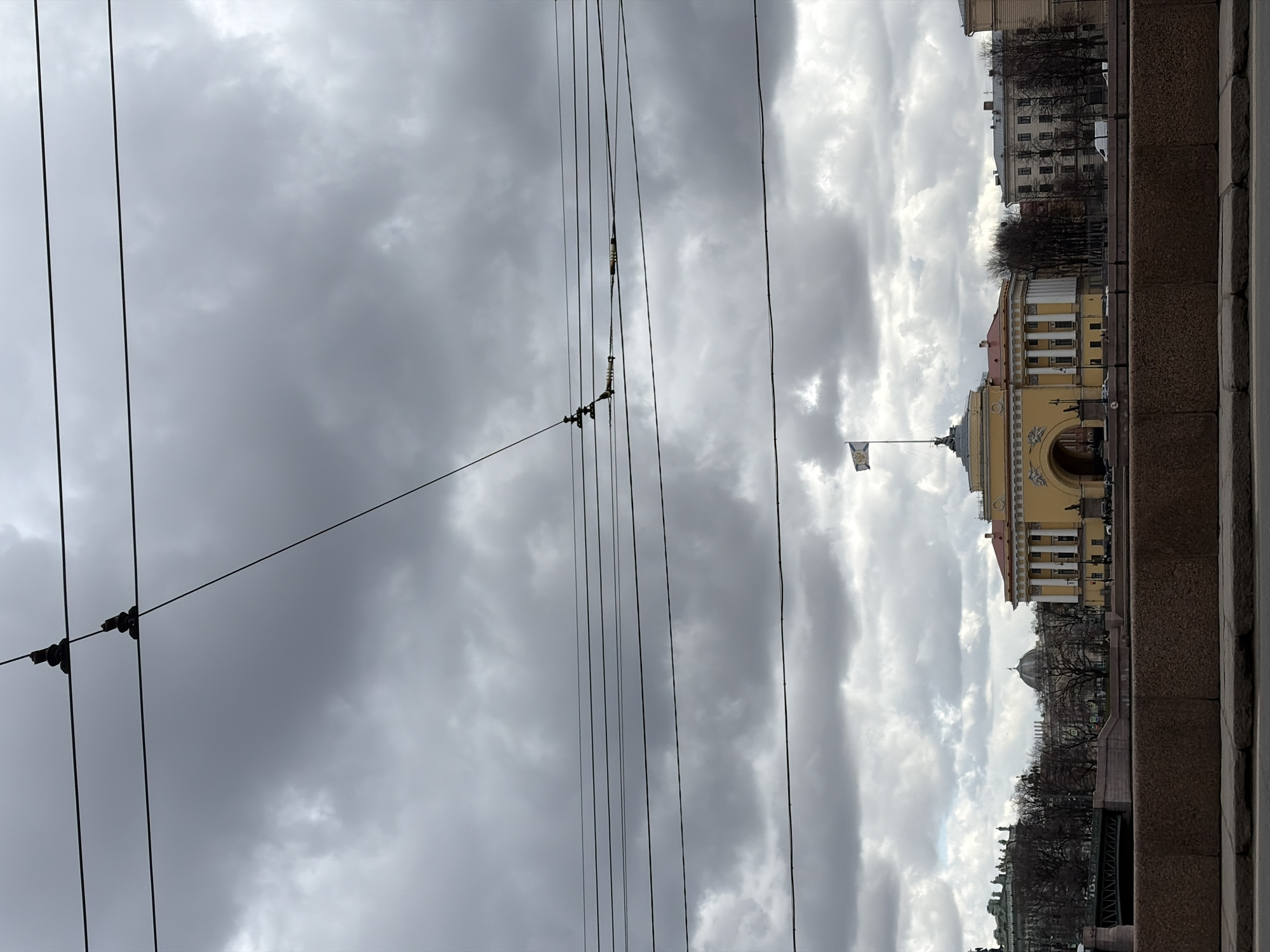 The image shows a view of the General Staff Building in Saint Petersburg, Russia, taken from across the canal. The foreground consists of a stone embankment and numerous overhead electrical wires. The General Staff Building is a yellow structure with a central arch and a flag flying on top. In the background, other buildings and trees are visible. The sky is overcast with dark clouds. The image has been rotated 90 degrees counterclockwise.