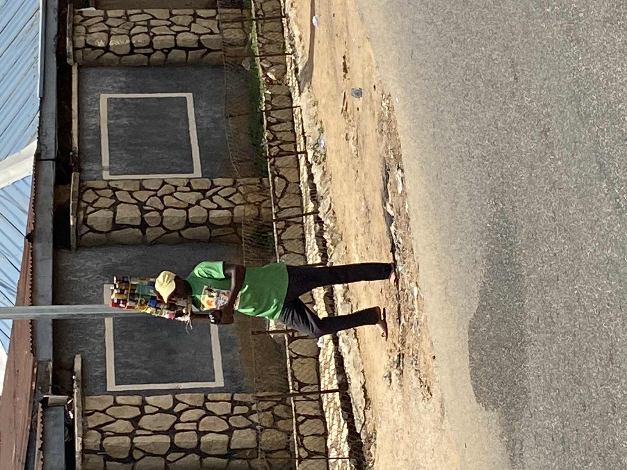 A male street vendor walks along a dusty path next to an asphalt road, in what appears to be a daytime scene under bright sunlight. He is wearing a bright green t-shirt, dark trousers, and a yellow baseball cap. He carries a rectangular tray filled with numerous small, colorful packaged goods, likely snacks, candies, or cigarettes, balanced on his head. His feet are bare.
To his left, the asphalt road stretches out, appearing dry and mostly clear. To his right, beyond the dirt path, stands a building with a distinctive architectural style. The lower part of the building's wall and frames around two prominent rectangular sections are decorated with rough-hewn, light-colored stones, creating a textured pattern. The central parts of these rectangular sections and the upper wall are a darker, possibly grey or charcoal, color. A low, dark, metal fence runs parallel to the dirt path, separating it from the building. Further to the left, a corrugated metal roof is partially visible. A tall, slender grey pole stands in the foreground. The overall setting, including the street vending activity and building styles, is consistent with a location in Bukuru, Nigeria. No discernible text is visible on any objects or surfaces.
